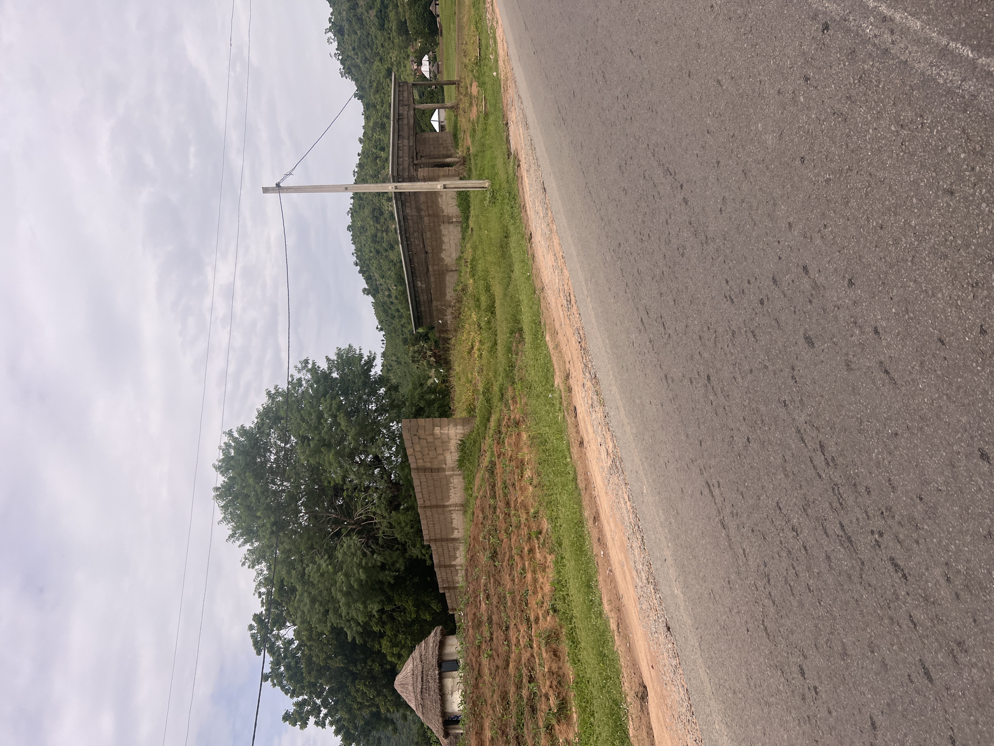 The image depicts a scene in Minda, Nigeria. The foreground features a paved road running diagonally across the frame, with a dirt shoulder and sparse vegetation. To the left of the road, a small, thatched-roof hut sits in a field of dry grass and young plants. Behind the hut, a wall constructed of concrete blocks separates the field from an area with lush green trees. Further in the background, there are more concrete block structures, possibly under construction or unfinished, and a densely forested hillside. Overhead, a cloudy sky dominates the upper portion of the image, with power lines stretching across it. The lighting suggests it is daytime, possibly overcast. The overall impression is of a rural or semi-rural environment with developing infrastructure. There are no visible people or text in the image.