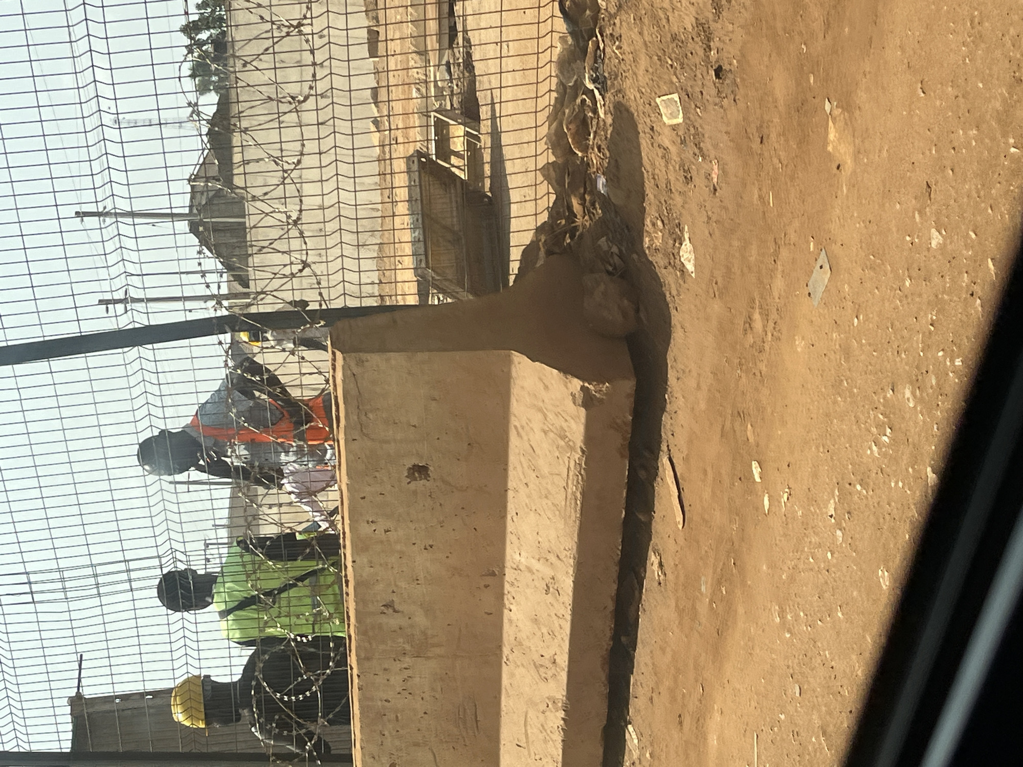 The image displays an outdoor scene, likely a construction site or an area undergoing development, set in Keffi, Nigeria. The photograph is taken from a slightly elevated position, looking down towards a dirt ground and a fence.
In the foreground, a rough dirt ground occupies the right side, showing varying shades of brown earth with small pebbles and some scattered debris, including what appears to be plastic or paper scraps. On the left side, a sturdy concrete barrier or wall runs horizontally, separating the immediate foreground from an area beyond a metal mesh fence.
Behind this fence, several individuals are visible, likely workers. Two figures are prominent: one wears a bright orange safety vest and a dark hard hat, while another wears a neon green safety vest and a dark hard hat. A third individual with a yellow hard hat is partially visible further down. They appear to be standing or engaged in tasks behind the fence, which is topped with coiled barbed wire, suggesting a secured area. The fence itself is made of interwoven metal wires forming a grid pattern.
The scene is brightly lit by natural sunlight, casting distinct shadows from the fence and the concrete structures, indicating a clear, sunny day, possibly morning or late afternoon given the angle of the light. No discernible text is visible on any signs or objects within the frame. The overall impression is one of an active work zone in a dry, open environment.