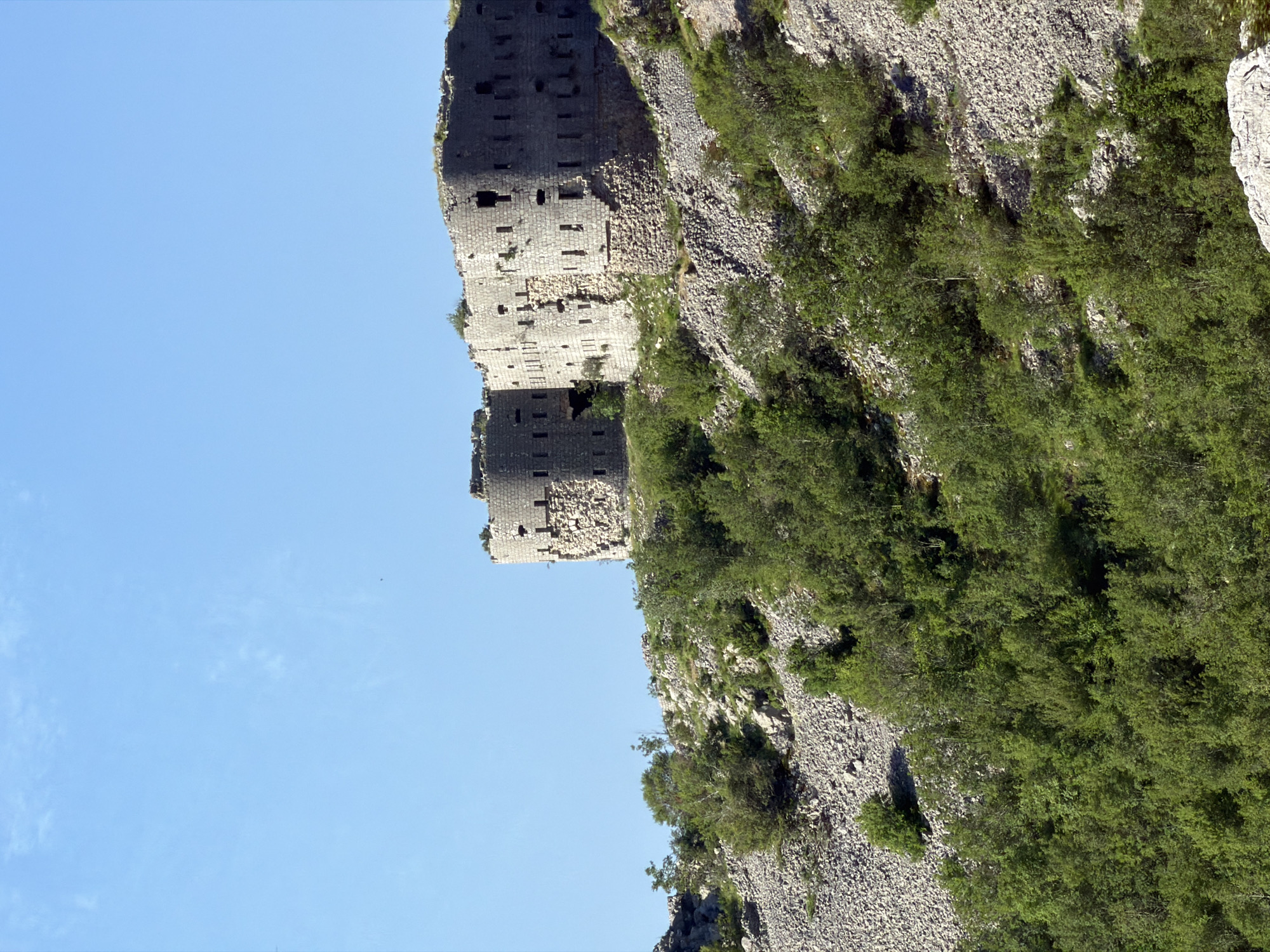 The image shows the ruins of a fortress perched on a steep, rocky hillside covered in sparse vegetation and trees. The sky above is clear and blue with a few wispy clouds. The fortress itself is constructed from stone blocks and features multiple round towers and walls with numerous openings, suggesting it was once a defensive structure. Some sections of the fortress appear to be in disrepair, with visible damage and crumbling stonework. The overall impression is one of an ancient, abandoned structure in a rugged natural setting. The location is identified as Stanišići, Montenegro.