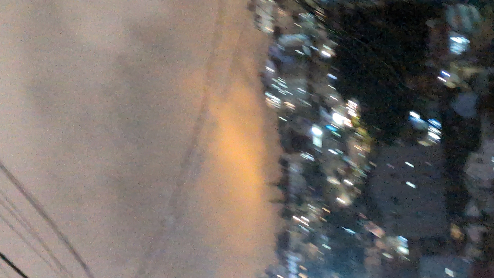 The image depicts a blurry, high-angle view of a city at dusk or night, likely Da Lat, Vietnam, given the presence of telephone or power lines in the foreground. The sky is overcast with a prominent band of orange-yellow light near the horizon, suggesting sunset or urban light pollution interacting with clouds. Below the sky, the indistinct forms of buildings and trees are visible, illuminated by numerous small, blurred lights, indicative of a populated urban area. The overall impression is one of a city coming to life with artificial light as natural light fades.