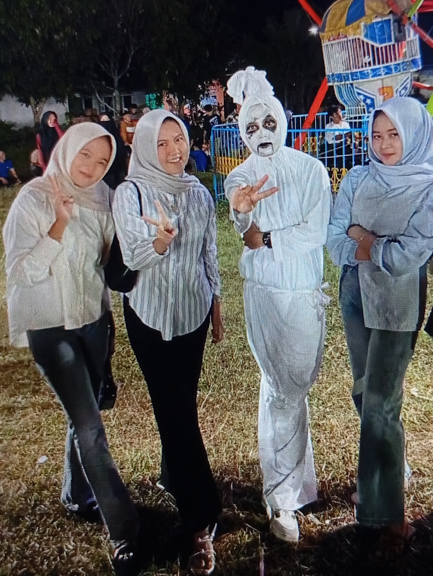 The image shows four people standing in a grassy area, possibly a park or an amusement area, at night. Two women are on the left, wearing headscarves and casual clothing. The woman on the far left wears a cream-colored, loose-fitting top and dark pants. She is making a peace sign with her right hand. The woman next to her wears a striped long-sleeved shirt and dark pants, also making a peace sign with her right hand.
In the center, a person in a ghost costume stands with their face painted white with black accents around the eyes and mouth, resembling a spooky apparition. They are wearing a white, full-body suit, and their head is covered with white fabric. This person is also making a peace sign with their right hand.
On the far right, another woman wearing a light blue headscarf and a light blue, long-sleeved top stands with her arms crossed. She is wearing dark pants.
In the background, there are trees and the illuminated structures of what appears to be a carnival or amusement park, including a Ferris wheel-like ride with colorful lights. The ground is covered with dry grass and some scattered leaves. The presence of the amusement park rides and the nighttime setting suggest this scene might be at an event in Lhokseumawe, Indonesia, known for its cultural festivals and community gatherings that sometimes include amusement attractions.