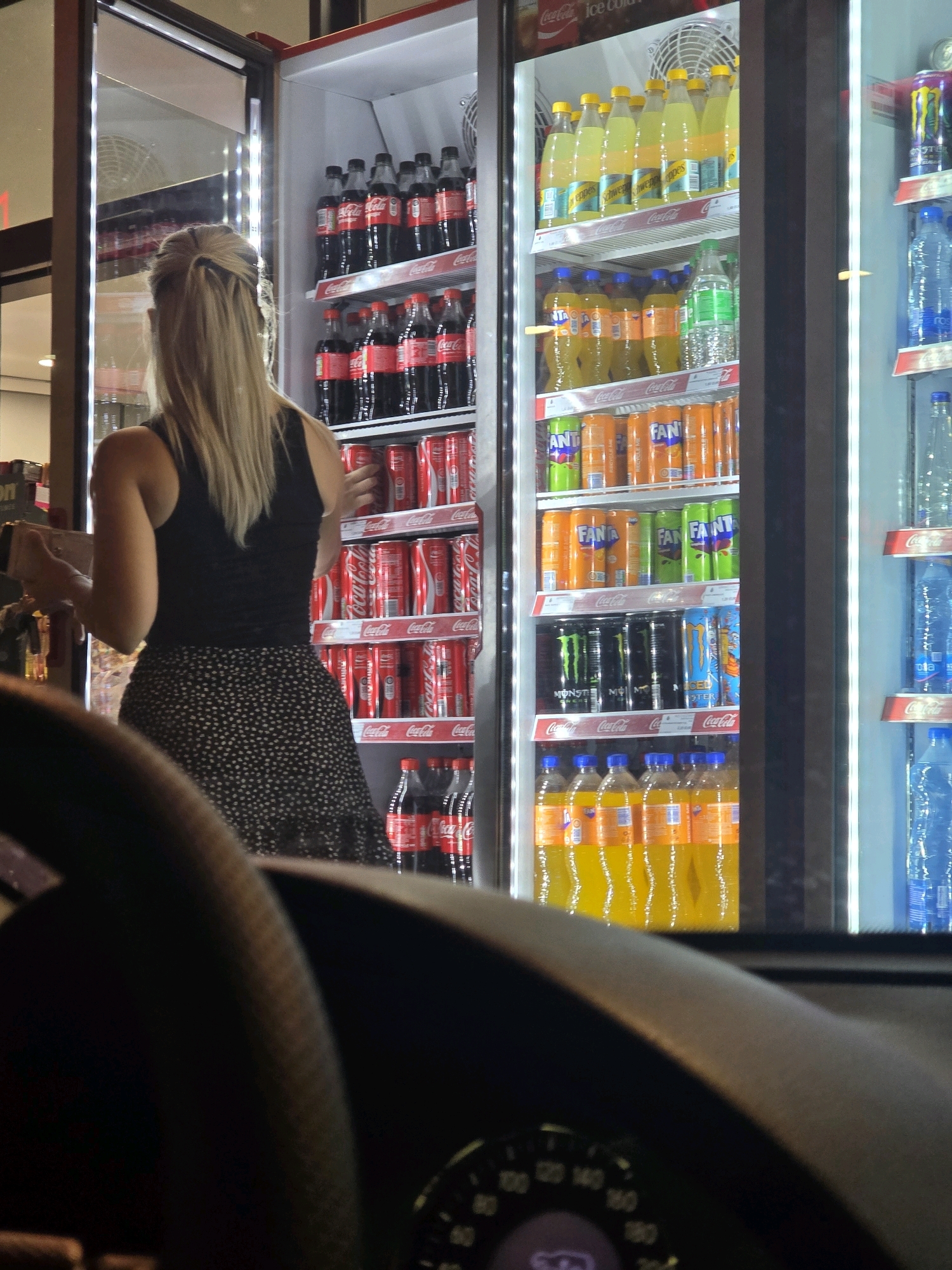 A woman with blonde hair tied in a ponytail stands in front of a brightly lit, open refrigerator at what appears to be a convenience store. She is wearing a black tank top and a dark skirt with a white polka dot pattern. Her back is to the camera, and she is reaching into the refrigerator. The refrigerator is stocked with numerous bottles and cans of various beverages, including Coca-Cola, Fanta, and energy drinks. The overall lighting suggests it is nighttime or the store is dimly lit. The image is captured from a low-angle perspective, possibly from inside a vehicle, as the lower portion of the frame shows a car's dashboard and steering wheel. The scene appears to be in Podgorica, Montenegro, as indicated by the location tag.