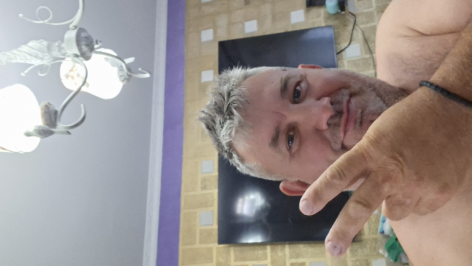 A middle-aged Caucasian man with graying, spiky hair and a beard smiles into the camera, making a peace sign with his right hand. He is bare-chested. The background includes a large flat-screen television, a patterned wallpaper, and a ceiling with a chandelier. A section of purple-painted wall is visible above the wallpaper. The location of Voinova, Moldova is not discernible from the image.