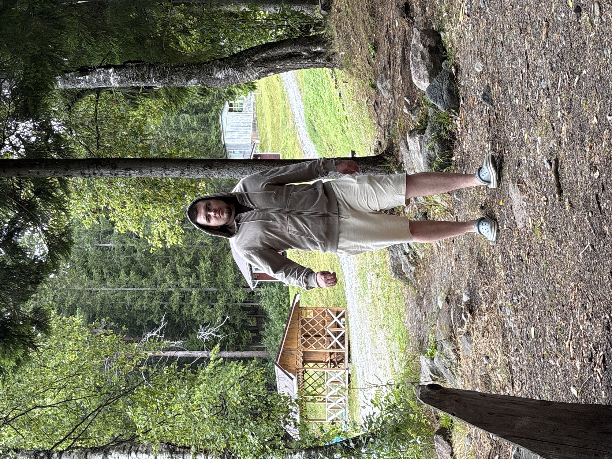 A young Caucasian man stands on a gravel path in a wooded area. He is wearing a light brown zippered hoodie with the hood up, and light beige shorts. He has a serious expression on his face and is looking directly at the camera. In the background, there are several wooden structures, including a gazebo with intricate lattice work, and a small building with light blue siding. The area is surrounded by tall trees with dense green foliage. The ground is covered with gravel, dirt, and fallen leaves. The overall setting appears to be a park or campground.