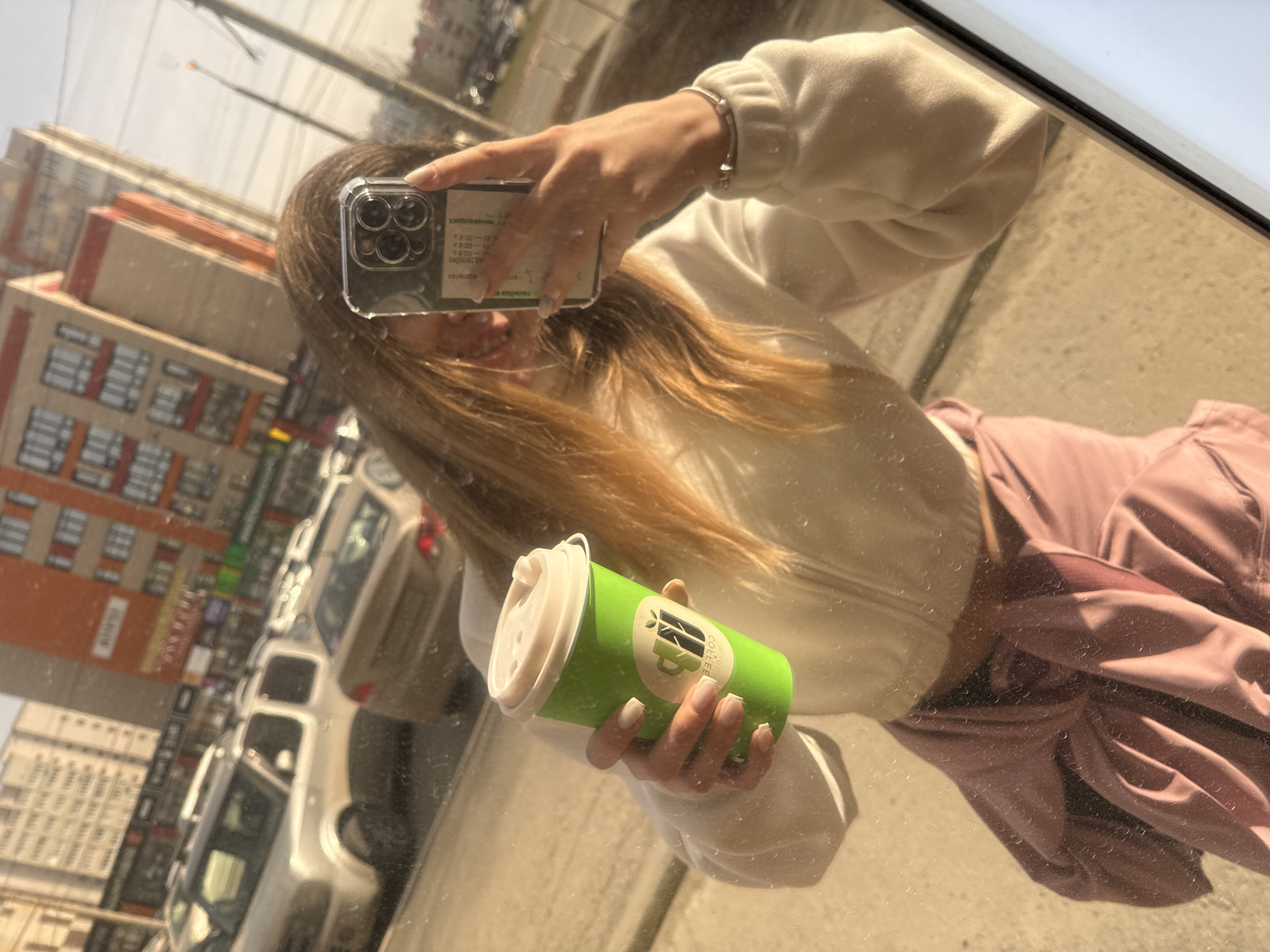 A woman takes a mirror selfie outdoors, holding a green disposable coffee cup in her left hand and a smartphone in her right. The smartphone has a clear case with triple cameras. The woman has long, light brown hair and wears a white zip-up sweater and a pink skirt or pants. The reflection in the mirror shows buildings and parked cars, suggesting an urban environment. The coffee cup has text and a logo that includes "MOOD COLLEE" with other indecipherable text and design. The clear case of the smartphone has a label or information sheet underneath, with text including "KasbreesiNG" and times "0800-12" and "08:00-15." There are also brand names such as "KBONHILL," "NOOTOTEX," and "KRONINGE" visible on the clear case. The background reflects city elements like buildings and streets.