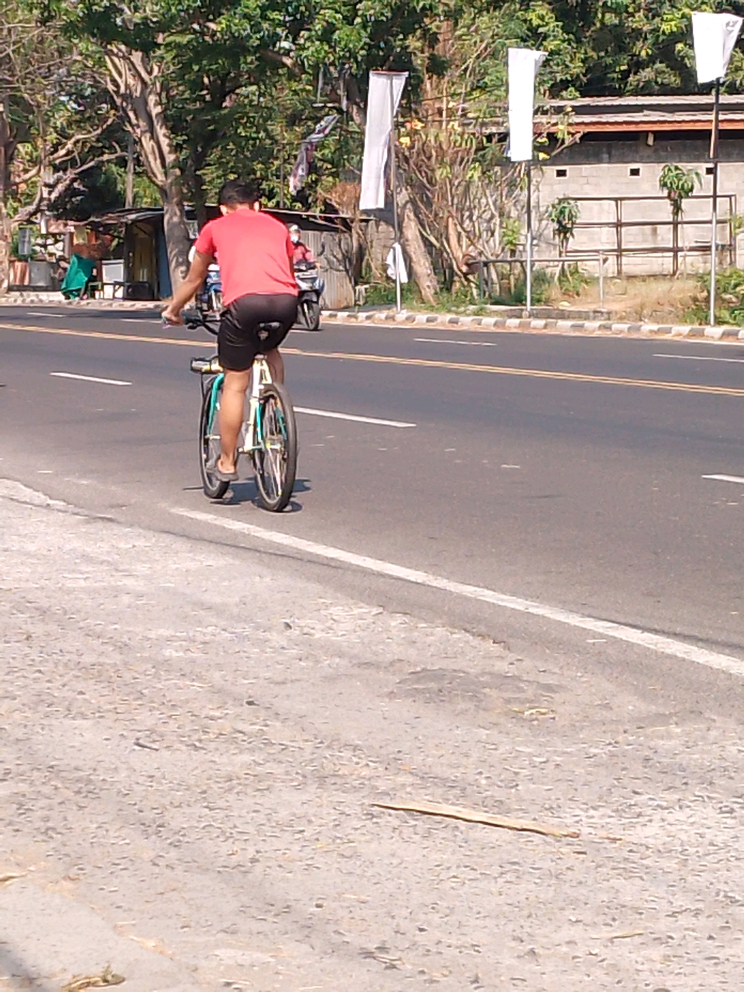 The image captures a person riding a bicycle on a street in Kutorejo, Indonesia. The rider, seen from behind, wears a red t-shirt and black shorts. The bicycle is a typical road bike with two wheels, a chain, and handlebars. The street is paved with asphalt and marked with white and yellow lines. On the side of the street, there are trees, buildings, and a few white flags. The scene appears to be a sunny day with clear weather.