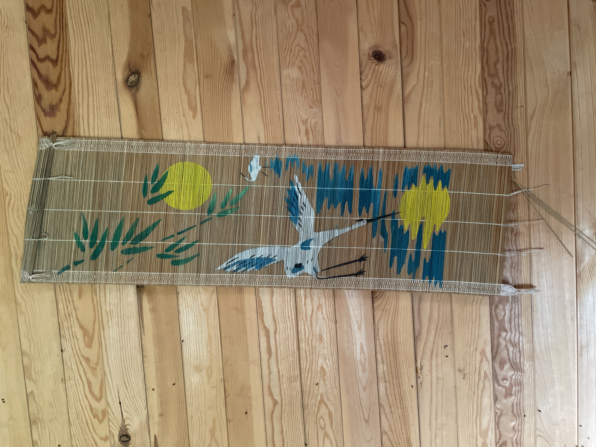 A woven bamboo mat with a painted Asian-inspired design lies horizontally on a light-colored wooden floor. The mat depicts a scene featuring a large white crane in flight, its wings spread. To the left of the crane, there are green bamboo leaves and a yellow sun. To the right, there are abstract blue and yellow shapes that could represent stylized mountains or water. The crane has black legs and a black beak. Two small white birds are flying near the top left of the mat. The mat is held together by thin white threads. Some of these threads are loose on the right side of the mat, suggesting it might be in the process of being made or is slightly damaged. The wooden floor has a prominent grain pattern and several knots, indicating it's likely made of natural wood planks. The lighting suggests it could be daytime, possibly indoors given the smooth wooden surface. There is no visible text in the image.
