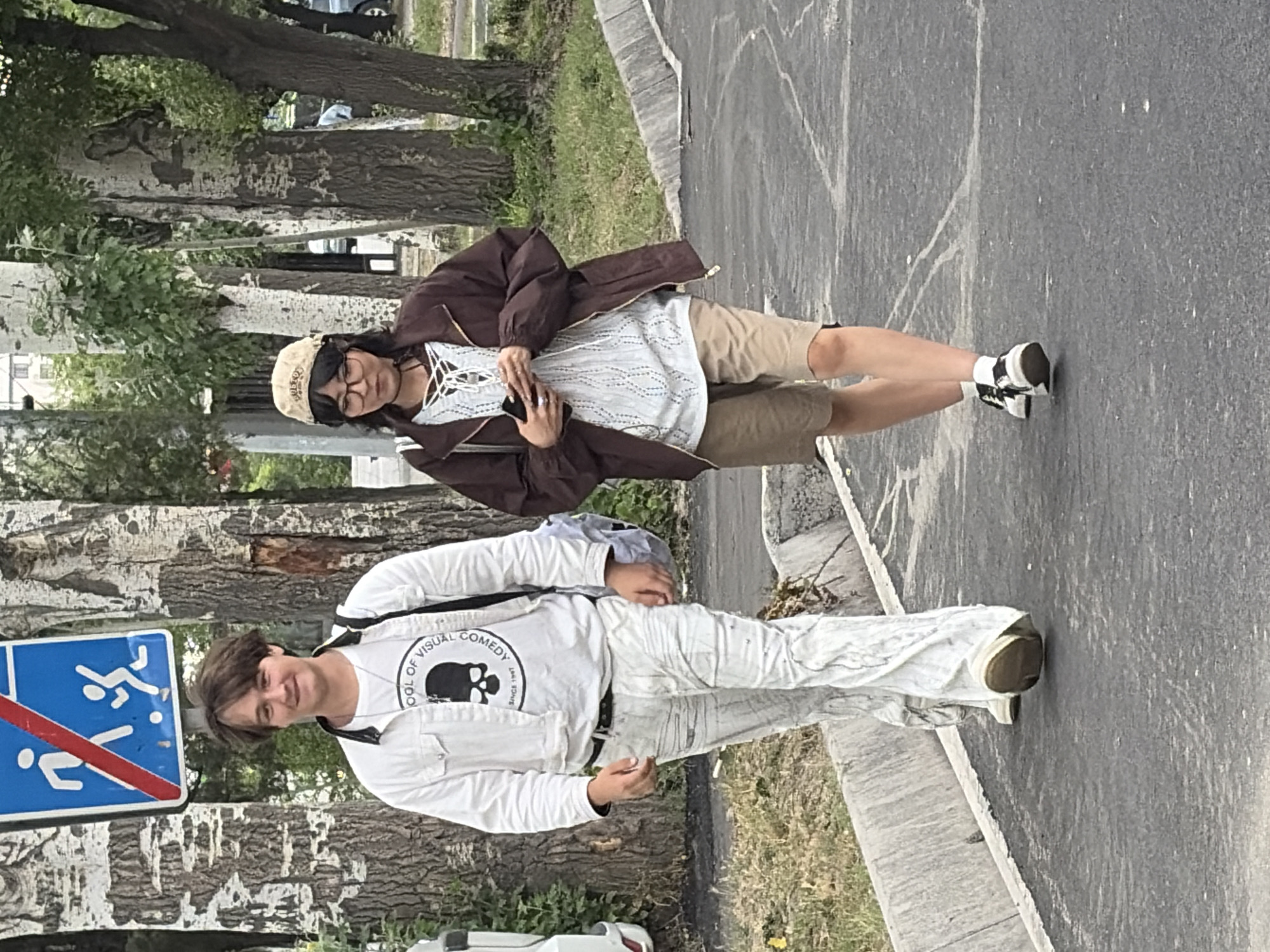 Two individuals, a male and a female, are walking on a paved path next to a grassy area and a line of trees. The male, on the left, is wearing a white denim jacket over a white t-shirt with a skull graphic, white flared pants, and light-colored shoes. He has dark hair and is looking forward with a slight smile. The female, on the right, is wearing a brown jacket over a white patterned top with a lace-up detail, khaki shorts, and black and white sneakers. She has dark hair, glasses, and is looking down at her phone. She is also wearing a light-colored baseball cap. A blue and red pedestrian crossing sign is visible in the background behind the male. The lighting suggests it is daytime.