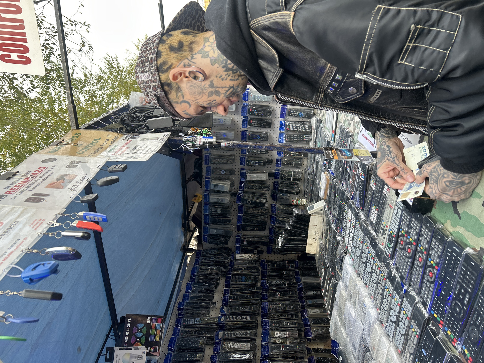 The photograph depicts a man with extensive facial and hand tattoos, wearing a black hooded jacket, browsing a street vendor's stall filled with an array of remote controls, presumably for sale. The man is holding what appears to be Argentinian pesos (200 notes) in his tattooed hand. The stall is covered with a blue tarp and features signs advertising replacement services for car alarm casings and automotive keys, as well as universal remote controls. It seems to be a market in Buenos Aires, Argentina as pesos in the photo display "200 Pesos".