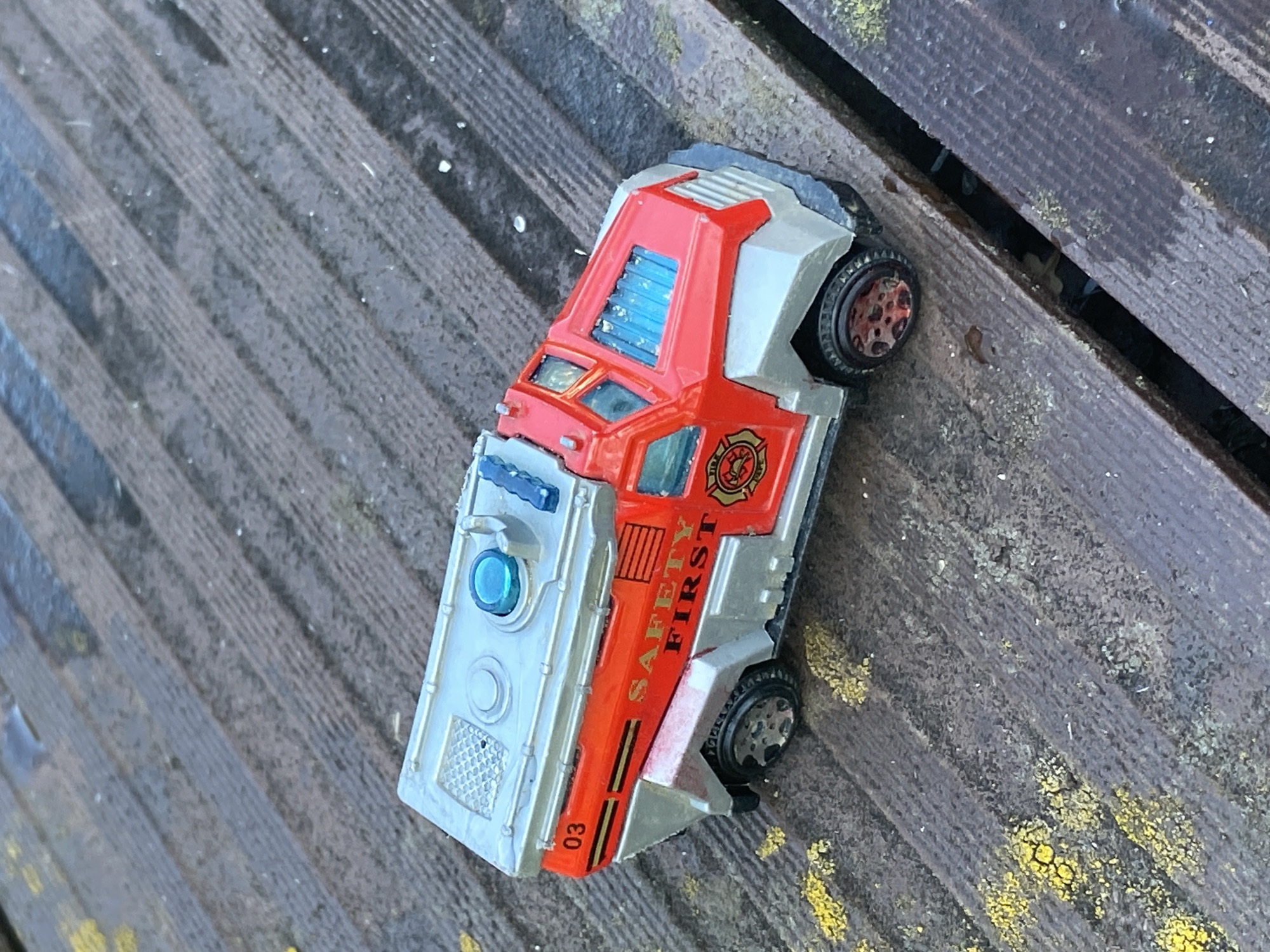 A red and silver toy fire truck with the words "SAFETY FIRST" printed on its side rests on a weathered wooden surface. The surface is made of planks with visible grain and is partially covered in yellow lichen and dark, wet patches. The toy fire truck has a blue light on its roof and a circular fire department emblem on its side. The number "03" is visible on the rear corner of the truck.
