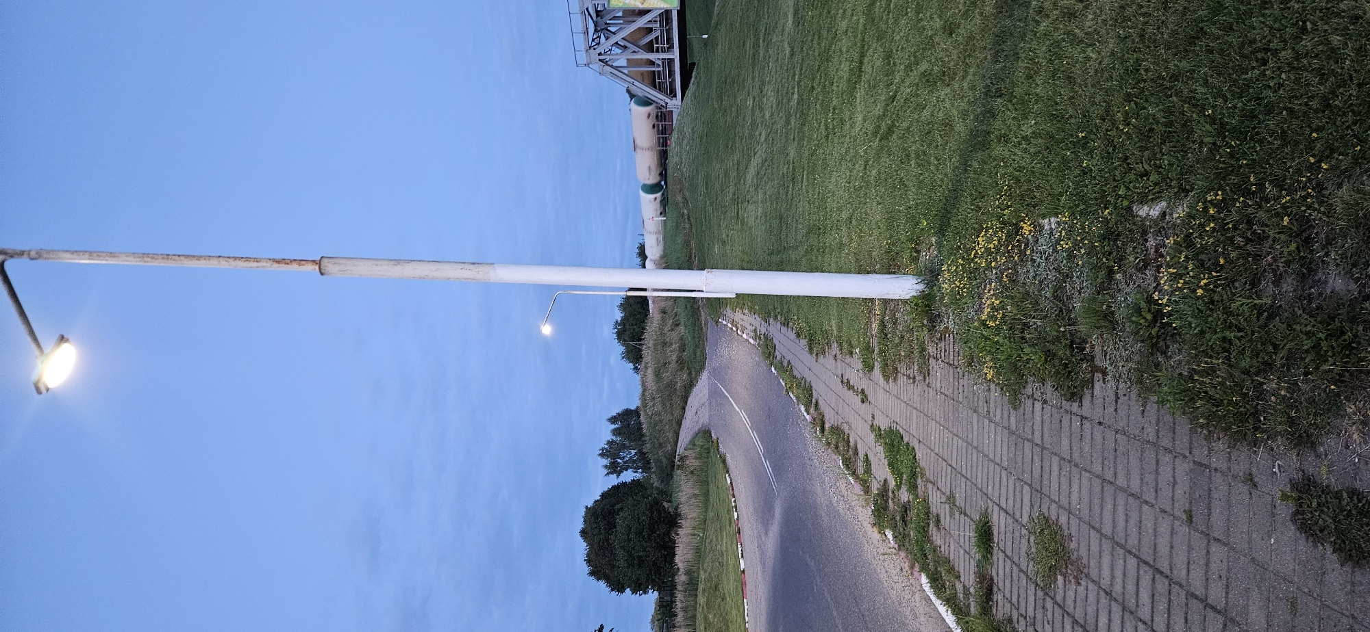 The image depicts a street scene at dusk, illuminated by a street lamp. The setting is the city of Vitsebsk, Belarus, specifically near a railway bridge. A paved road with white lane markings curves to the left, bordered by a sidewalk made of interlocking concrete pavers. Weeds and small yellow flowers grow along the edges of the sidewalk and road. To the right of the sidewalk, a grassy embankment slopes upwards towards a railway bridge, which supports a train of white tank cars. The sky is a deep blue with scattered clouds. A tall, white street lamp post stands prominently in the foreground, its light casting a glow. The overall impression is one of a quiet, rural-industrial intersection under twilight.