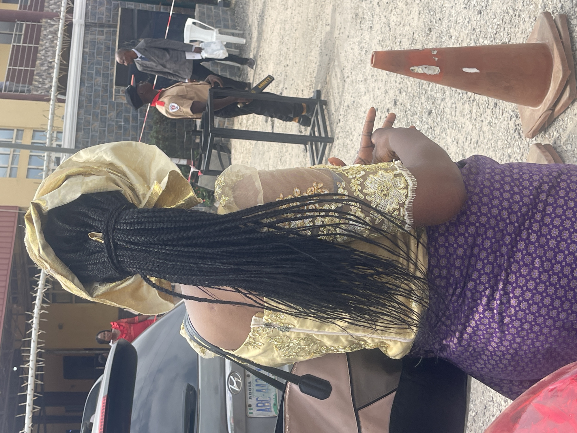 A medium shot depicts a scene outdoors, likely a checkpoint or entrance to a secured area, in Abuja, Nigeria. The main subject is the back of a woman with braided hair, wearing a gold-colored headwrap, and a gold-embroidered top over a purple patterned skirt. She is positioned next to a dark gray car, with the license plate number "ABC 442."
In the background, a uniformed security guard, possibly conducting security checks, stands behind a metal detector. Further back, there are people standing near a building, and a man in a suit is visible. A traffic cone is near the right side of the frame. The setting appears to be a paved area, potentially a parking lot or driveway. The lighting suggests it is daytime.