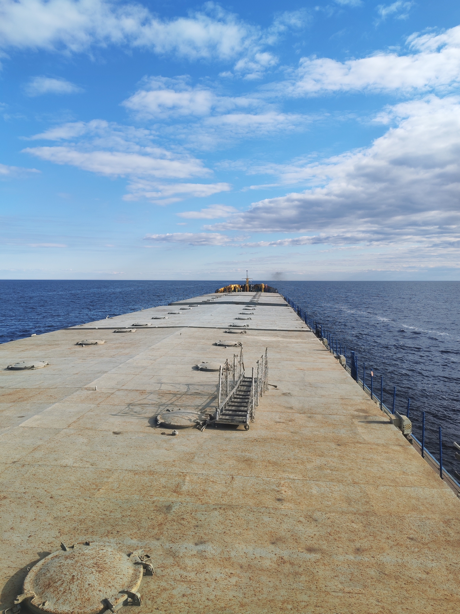 The image captures a wide-angle view from the stern of a large, flat-decked vessel, likely a barge or cargo ship, looking towards its bow as it navigates open water. The dominant foreground is the vessel's deck, a broad, rusty, light gray metal surface composed of large panels. Several round, reinforced metal hatches are prominently scattered across the deck, particularly in the lower left, exhibiting significant rust. Near the center-right of the deck, a set of portable metal stairs with handrails stands, casting a distinct shadow. Blue metal railings line the starboard side of the deck, separating it from the dark blue water that stretches to the horizon.
The water surface shows ripples and a faint wake trailing from the vessel on the right, indicating movement. The sky above is a bright, clear blue, adorned with scattered white cumulus clouds, with some larger, darker cloud formations visible towards the horizon on the right. In the far distance, towards the bow of the vessel, small yellow objects and structures, possibly equipment or containers, are visible. A few very small figures, seemingly wearing yellow safety gear, can be discerned near this distant equipment. Black smoke or exhaust appears to rise from the port side of the vessel's distant end. The scene depicts a clear, sunny daytime with fair weather, suggesting industrial activity or transport on a vast body of water. No discernible text is visible.