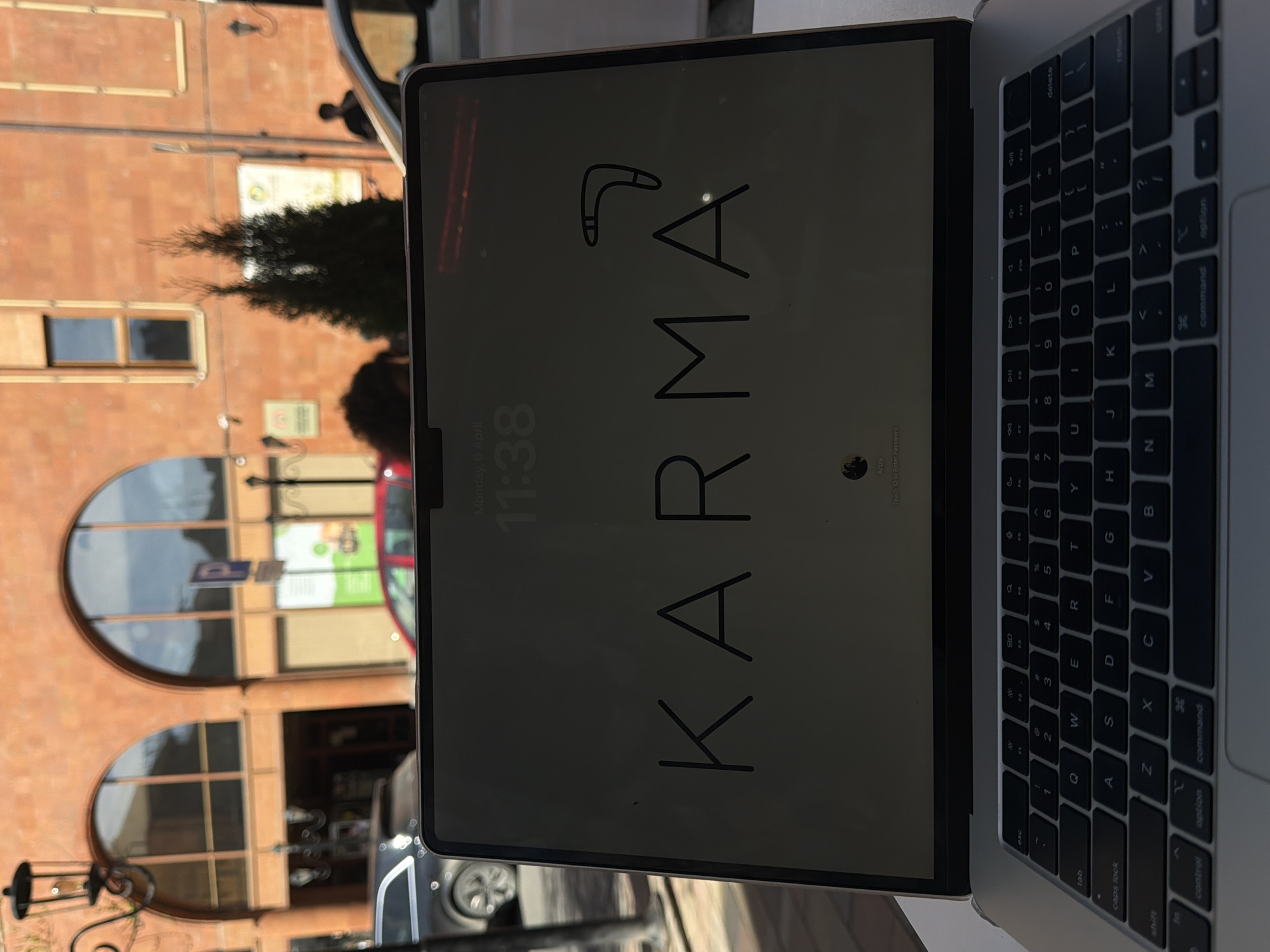 The image shows a close-up view of a MacBook screen displaying the word "KARMA" with a boomerang symbol above it. The screen also shows the time "11:38" and the date "Monday, 6 April". To the right of the screen, the keyboard of the MacBook is visible.
The background is out of focus and blurry, but the reflection in the screen provides some visual information. The reflection shows a building with arched windows and a tree. Based on the architectural style, this appears to be Yerevan, Armenia.