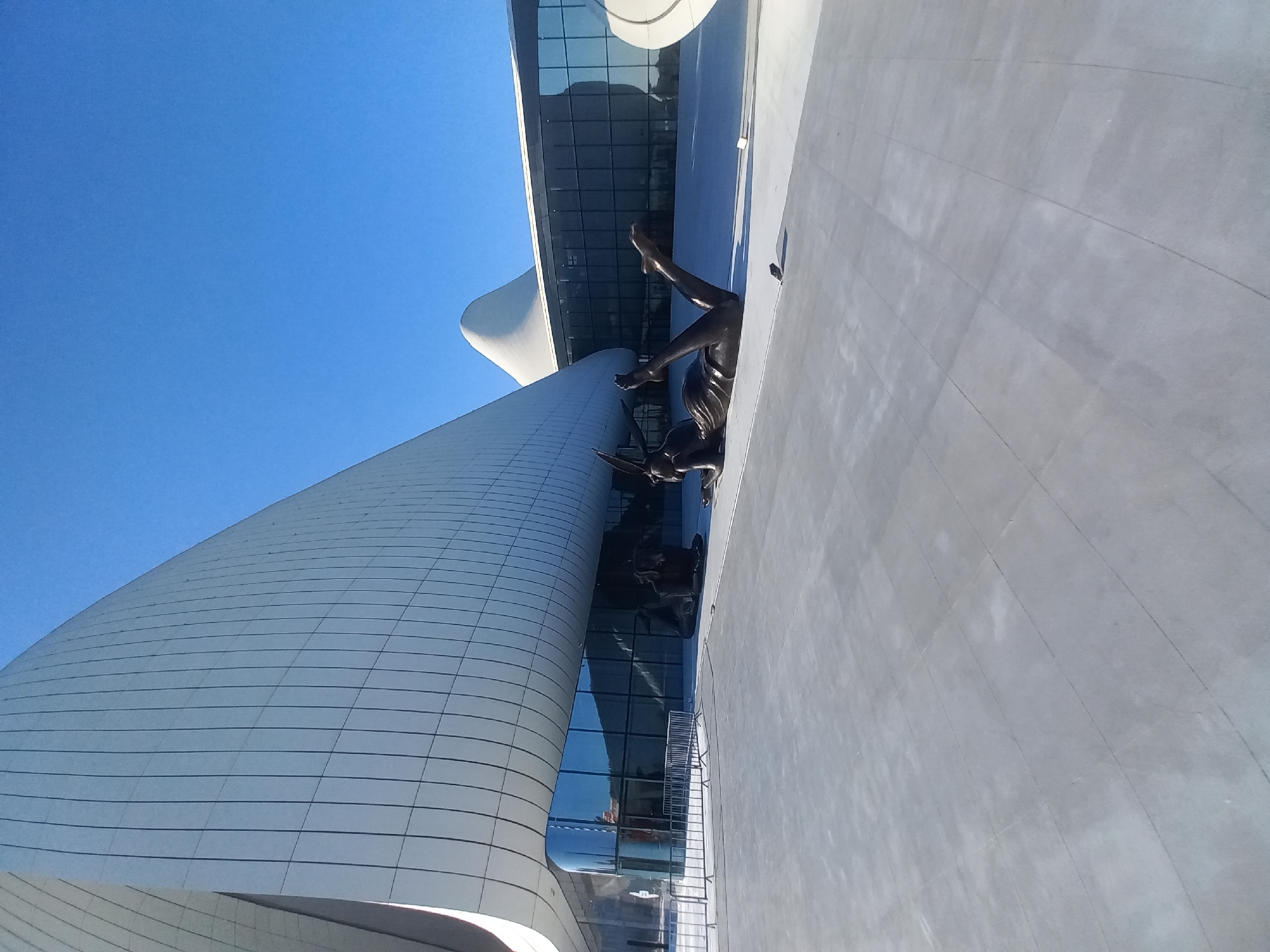 The image depicts the exterior of the Heydar Aliyev Center in Baku, Azerbaijan, a notable example of modern architecture characterized by its flowing, curved design. In the foreground, a bronze sculpture of a reclining nude figure is prominently displayed on the paved plaza. The sculpture is positioned to the right of center, facing left, with its legs extended upwards and its head resting on its arm. Behind the sculpture, the glass facade of the Heydar Aliyev Center reflects the clear blue sky and the building's distinctive white, curved roof. To the left of the sculpture, another dark bronze sculpture, appearing to be a seated figure or an animal, is partially visible. Metal barricades are set up on the plaza in front of the building's entrance. The overall scene is bathed in sunlight, casting shadows on the plaza.