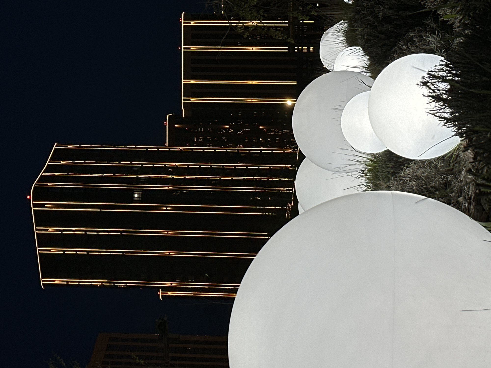 The image shows a nighttime scene featuring a cluster of illuminated white orbs in the foreground, with buildings visible in the background. The buildings are adorned with horizontal lines of light, suggesting a modern architectural style. The orbs appear to be decorative elements, possibly part of an outdoor installation. The sky is dark, indicating that the image was taken after sunset. The overall impression is that of an urban environment with a contemporary aesthetic. The location in the city of Ovoshchnoy, Russia, is not apparent based on the visual information in the image alone.