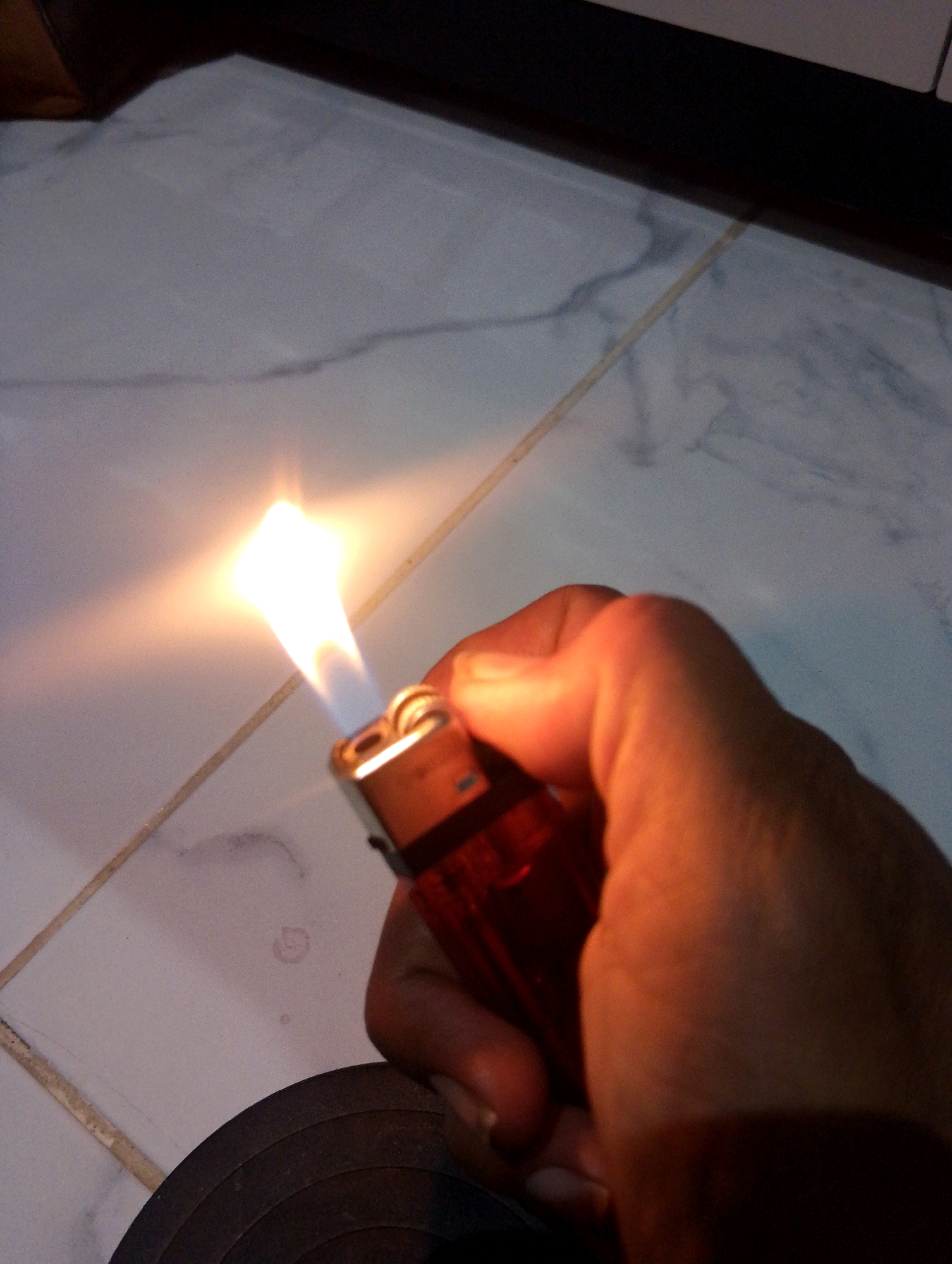 A person's hand holds a red lighter with a lit flame, illuminating the surrounding tiled floor. The floor appears to be white with grey marbling and is intersected by grout lines. A dark circular object, possibly a record or coaster, is visible in the lower left corner of the image. The lighting suggests a dimly lit environment. The image was taken in Sumbergirang, Indonesia.