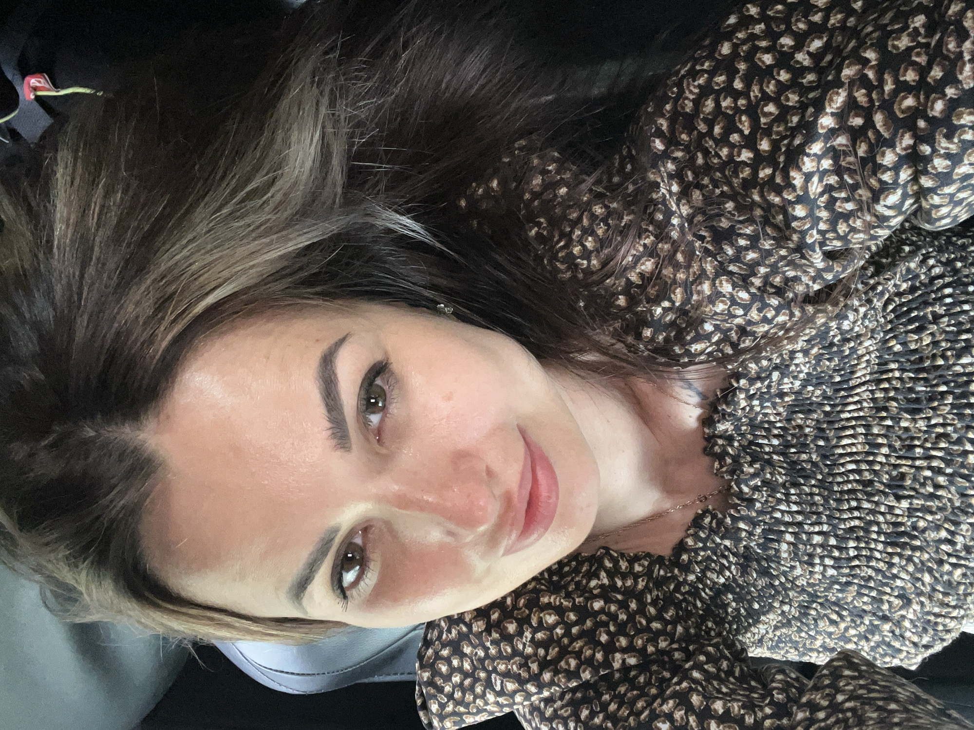 A young woman with brown hair and brown eyes is shown from the chest up, looking at the camera. She is wearing a dark brown and tan patterned blouse. Her hair is pulled back on the left side and falls over her right shoulder. The background is dark and out of focus, but appears to be the interior of a vehicle, possibly a car.