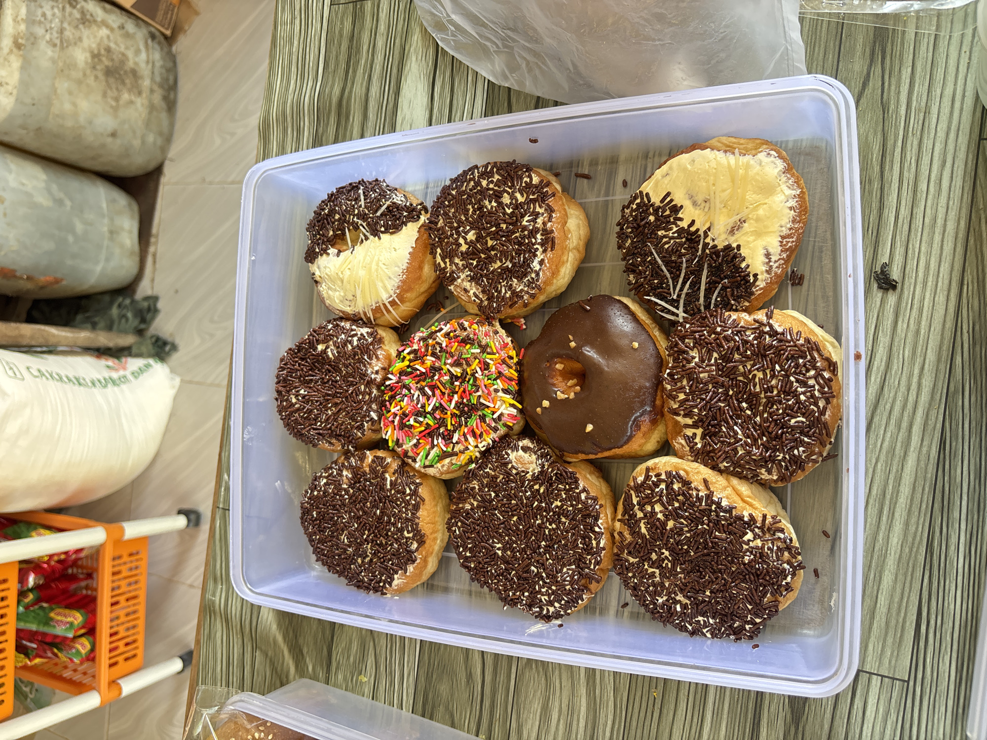 A transparent plastic container holds ten donuts on a wooden table. The donuts are arranged in three rows. The top row has three donuts: two covered in chocolate sprinkles and one with rainbow sprinkles. The middle row contains four donuts: two with chocolate sprinkles, one with a chocolate glaze, and one with white frosting and chocolate sprinkles. The bottom row features three donuts: two with chocolate sprinkles and one with white frosting and chocolate sprinkles. In the background, an orange shelf unit is visible, stocked with packets of snacks. To the right, two large white sacks are positioned behind the donuts, and further to the right, a clear plastic bag is draped over the edge of the table. The scene is indoors, likely in a food stall or shop. The image was taken in Lhokseumawe, Indonesia.