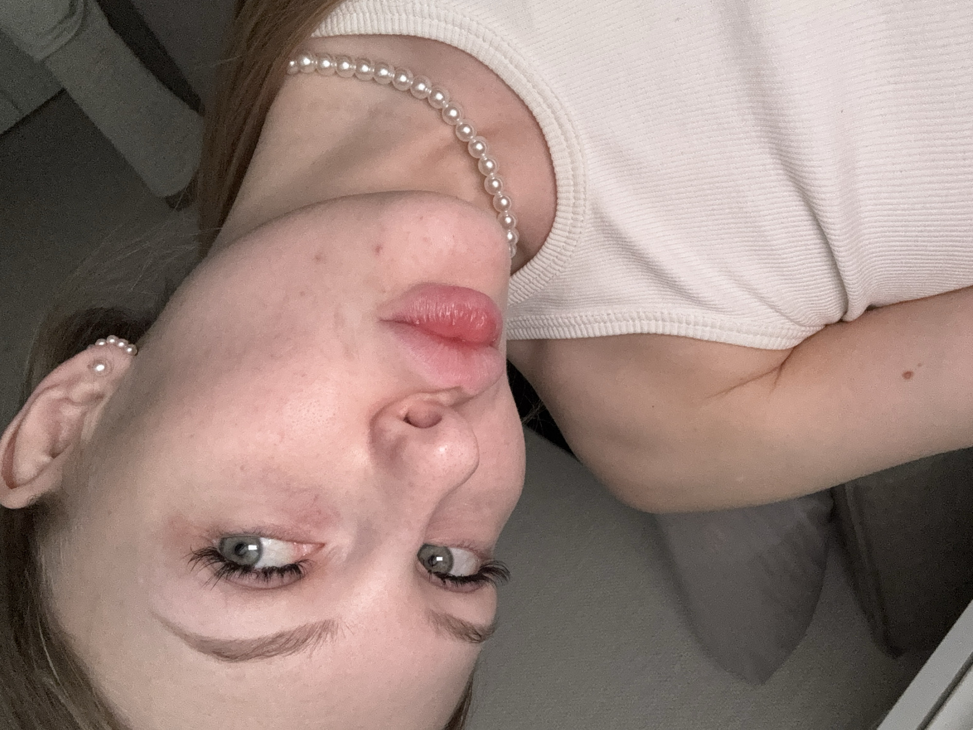 The image shows a close-up of a fair-skinned person from an unusual, upside-down perspective. The person is wearing a white ribbed tank top and a pearl necklace. The person's face is visible, including their eyes, nose, and lips, which are slightly pursed. An earring is visible in the left ear. The background is blurry and appears to be a room with furniture. The location, Perm, Russia, is not discernible from the image.