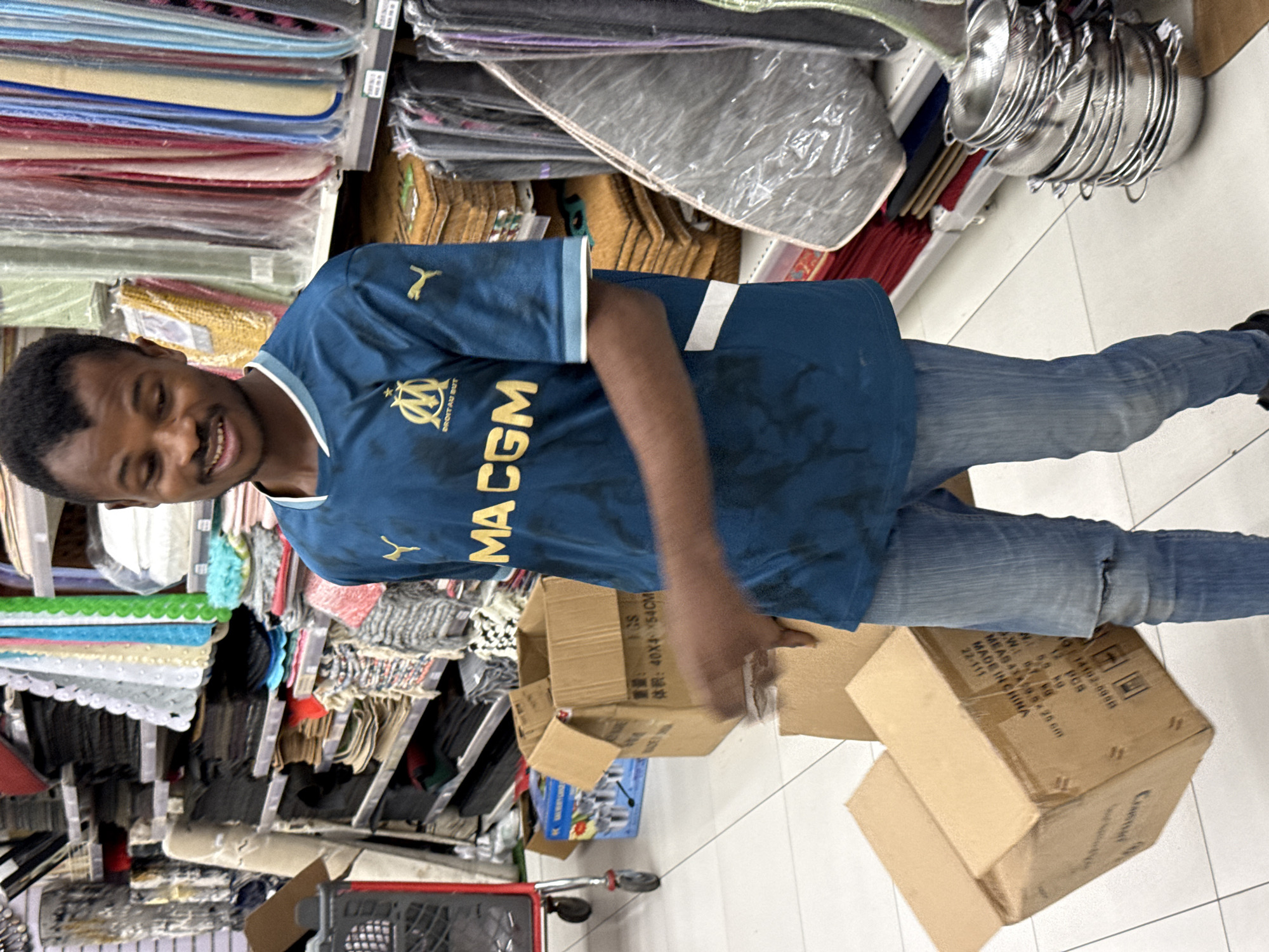 A man wearing a blue Marseille football jersey and jeans is standing in a store aisle next to shelves stocked with various home goods, including table cloths and kitchenware. He is holding and examining several stacked cardboard boxes. A shopping cart is visible near the bottom of the image. The packaging on one of the boxes indicates that it contains manufactured goods, with the text including the phrase "MADE IN CHINA". The image was likely taken indoors. It is not possible to confirm the location as Abuja, Nigeria, from the image.