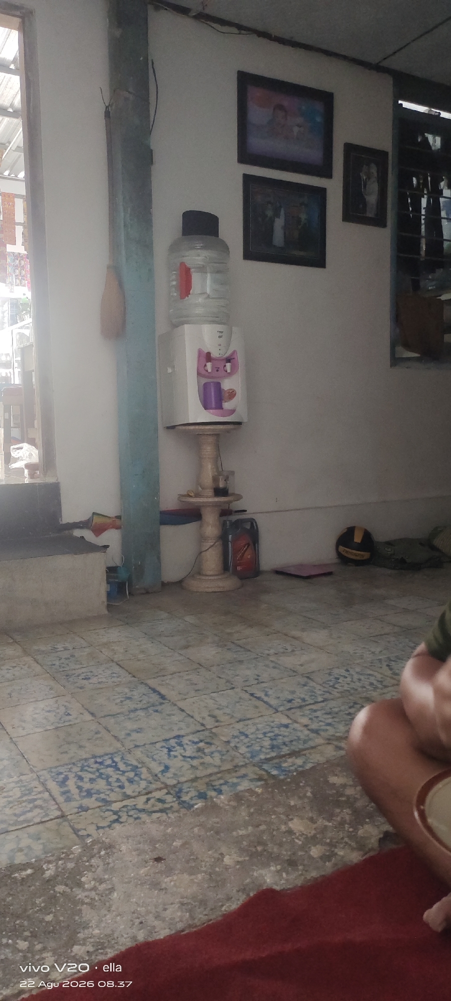 The image depicts an interior scene, likely a room in a residence in Simo, Indonesia. In the foreground, the floor is tiled with a pattern of blue and white squares. A portion of a person's legs and torso, wearing a green shirt, is visible in the lower right corner, and a red rug is spread out in the foreground.

The midground features a white water dispenser topped with a large clear bottle of water. Below the dispenser is a small, ornate stand with a glass containing a dark liquid. To the left of the dispenser is a light blue support column. On the wall behind the dispenser are three framed photographs.

The background shows a window with vertical blinds or louvers, allowing natural light to enter the room. A broom is leaning against the light blue column. Other objects visible include a black and yellow ball, a red container that appears to be a fuel can, and some green fabric. The walls are painted white. The overall impression is of a lived-in domestic space.