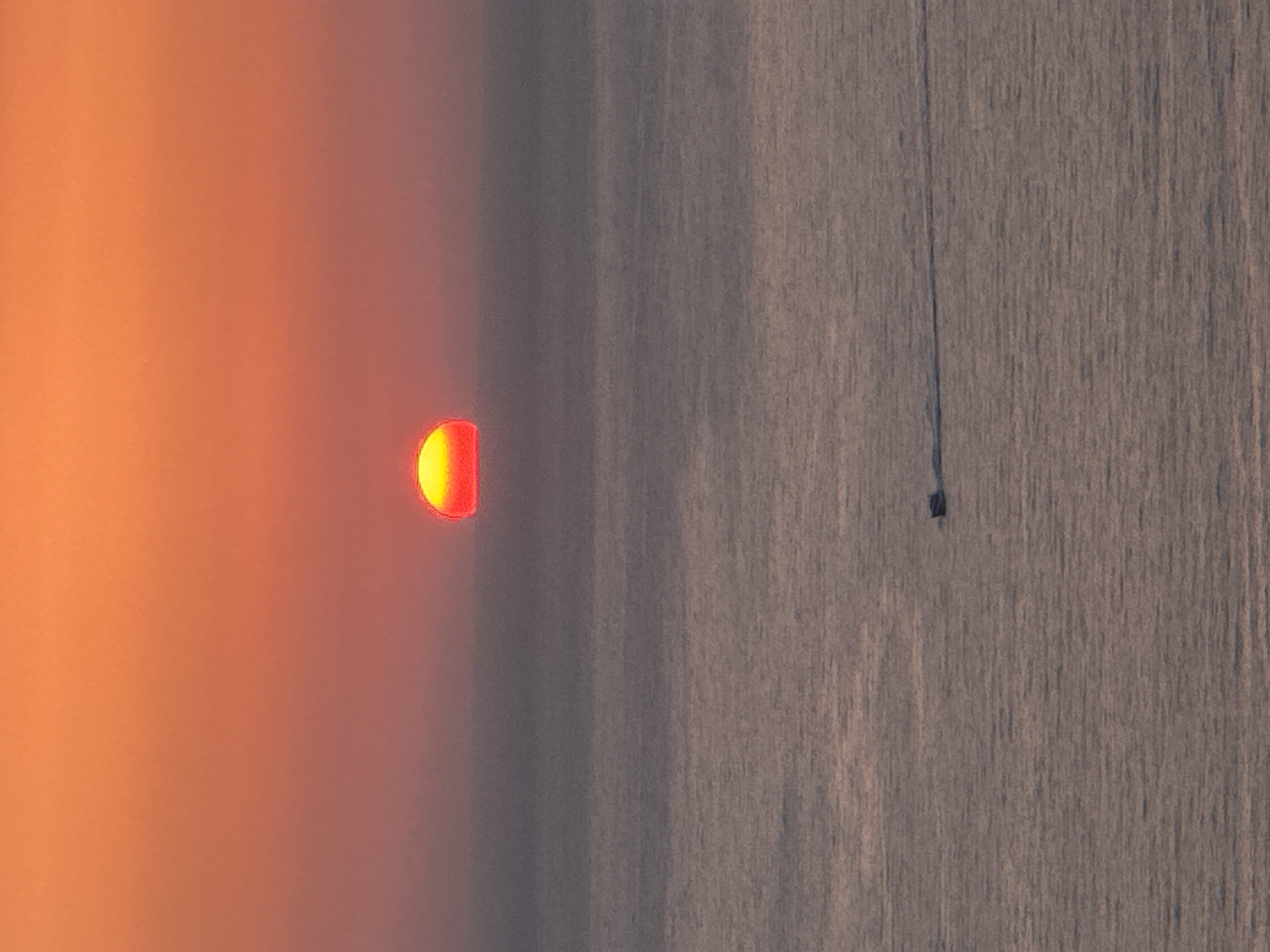 A sunset casts a vibrant orange and red glow across the sky, with the sun appearing as a semi-circle just above the horizon. Below the sky, the sea stretches out, its surface reflecting the warm colors of the sunset. A small boat with a dark hull is visible on the water, moving from left to right, leaving a faint wake behind it. The overall scene conveys a sense of tranquility and the beauty of nature. The city of Zagora, Montenegro is not depicted in this image.