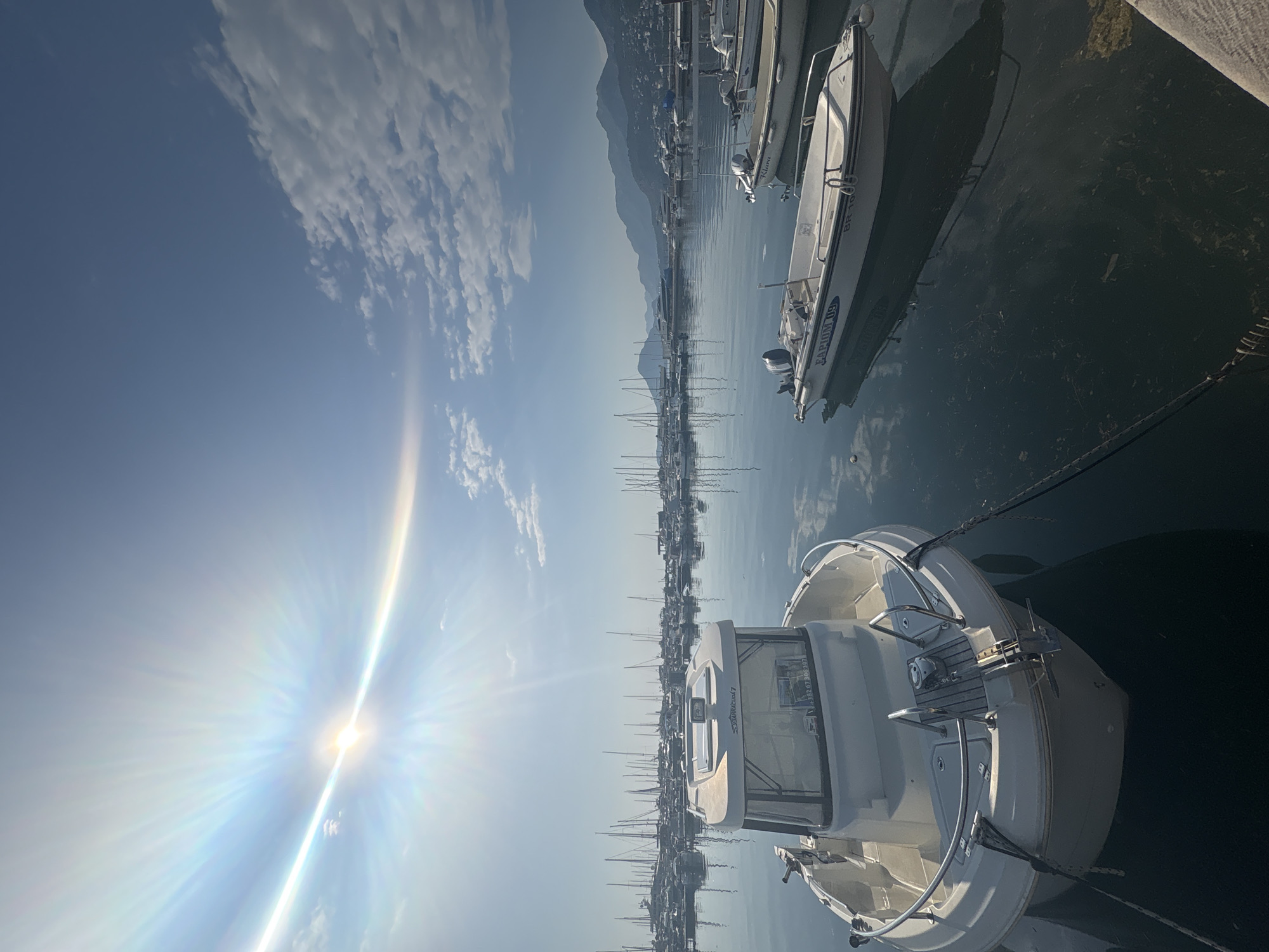 The image captures a sunny day at a marina in the city of Bar, Montenegro. The sky is blue with scattered white clouds, and the sun shines brightly, causing lens flare. In the foreground, a white motorboat is docked, with its stern facing the viewer. The boat is secured with ropes. In the background, there are numerous sailboats and other boats moored in the marina. Beyond the marina, there is a view of mountains.