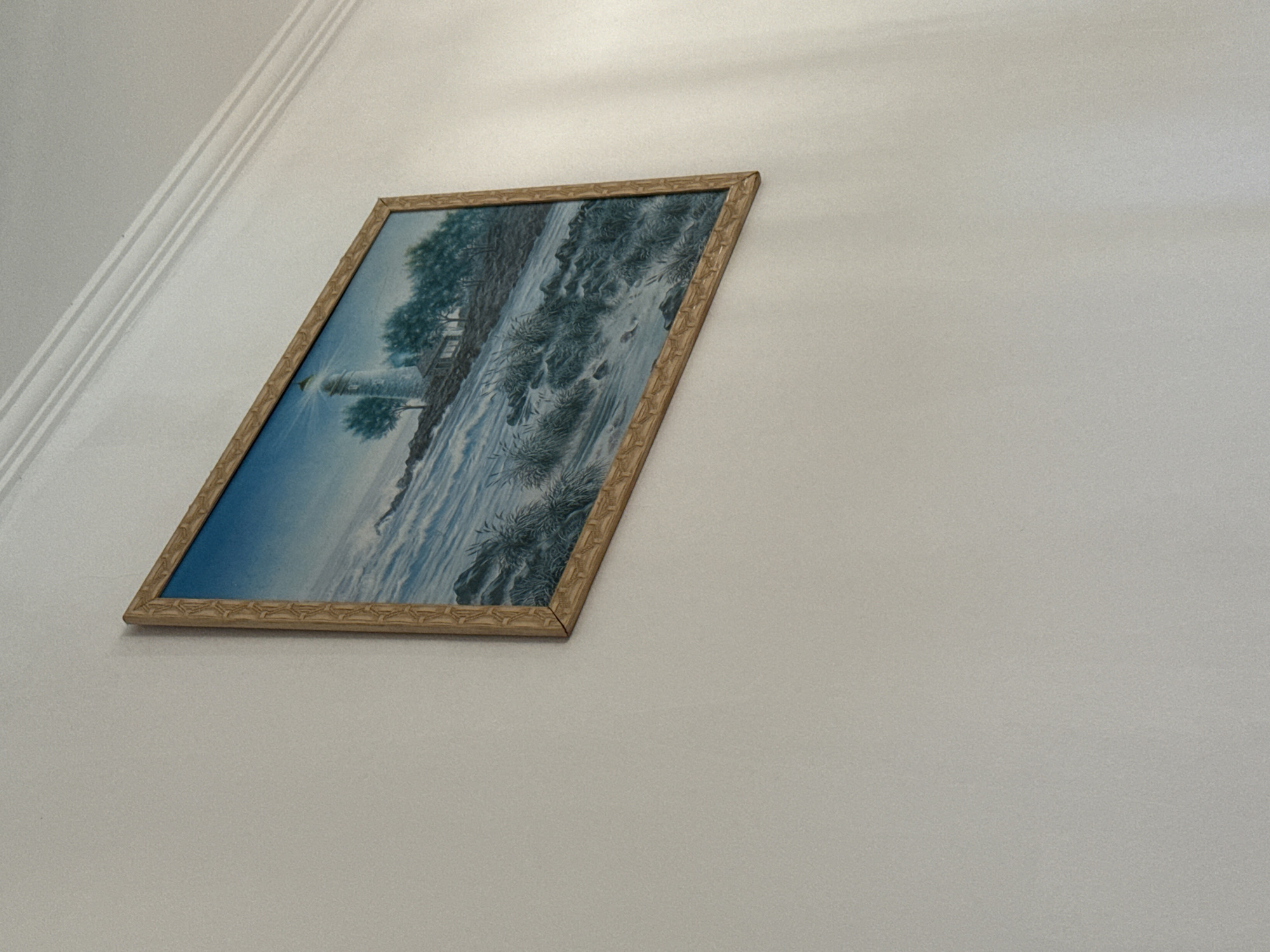 A framed painting of a lighthouse and trees on a rocky shore is hung on a white wall. The painting is in landscape orientation. The wall has subtle horizontal molding near the ceiling. The lighting suggests the scene is illuminated from the right. There are no people or animals visible in the image. The painting is positioned off-center and tilted slightly.
