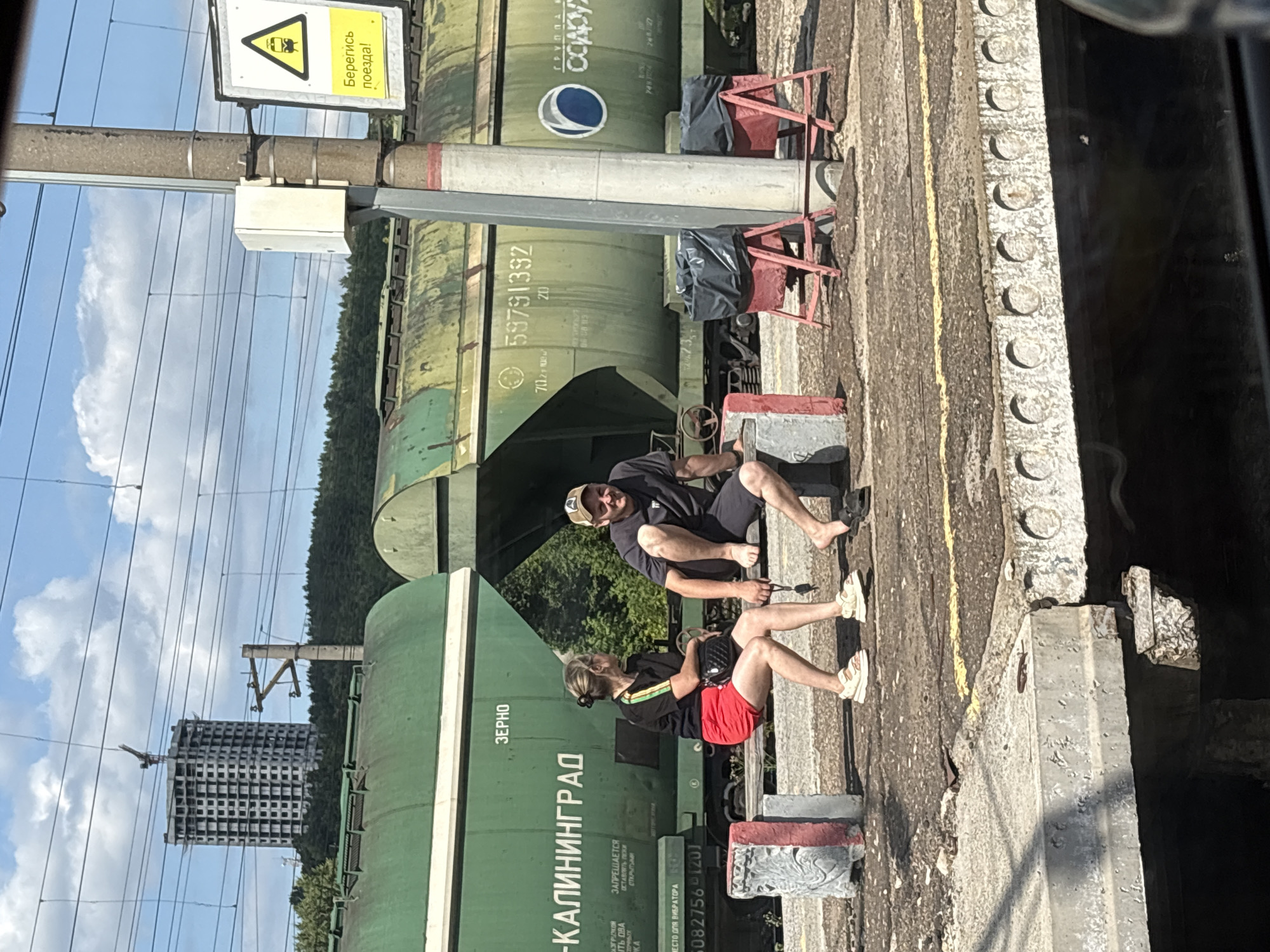 At a train station in Zelenodol'sk, Russia, two individuals are seated on a bench by a train track. On the left, a woman with blonde hair pulled back in a bun wears a black t-shirt with colored stripes and red shorts, with white sandals on her feet. She holds a dark purse in her lap. To her right, a man with a baseball cap on his head and a dark t-shirt and shorts is seated with his legs extended. Both appear to be waiting. Behind them, a long green train car, labeled "ЗЕРНО" (GRAIN) and "-КАЛИНИНГРАД", is parked on the tracks. Further in the background, a tall, unfinished concrete building is visible against a blue sky with scattered clouds, and a dense line of trees borders the horizon. A utility pole with a sign warning to "Beware of the train!" stands to the right of the individuals, with two red trash receptacles with black bags placed at its base. The ground is a weathered concrete platform.