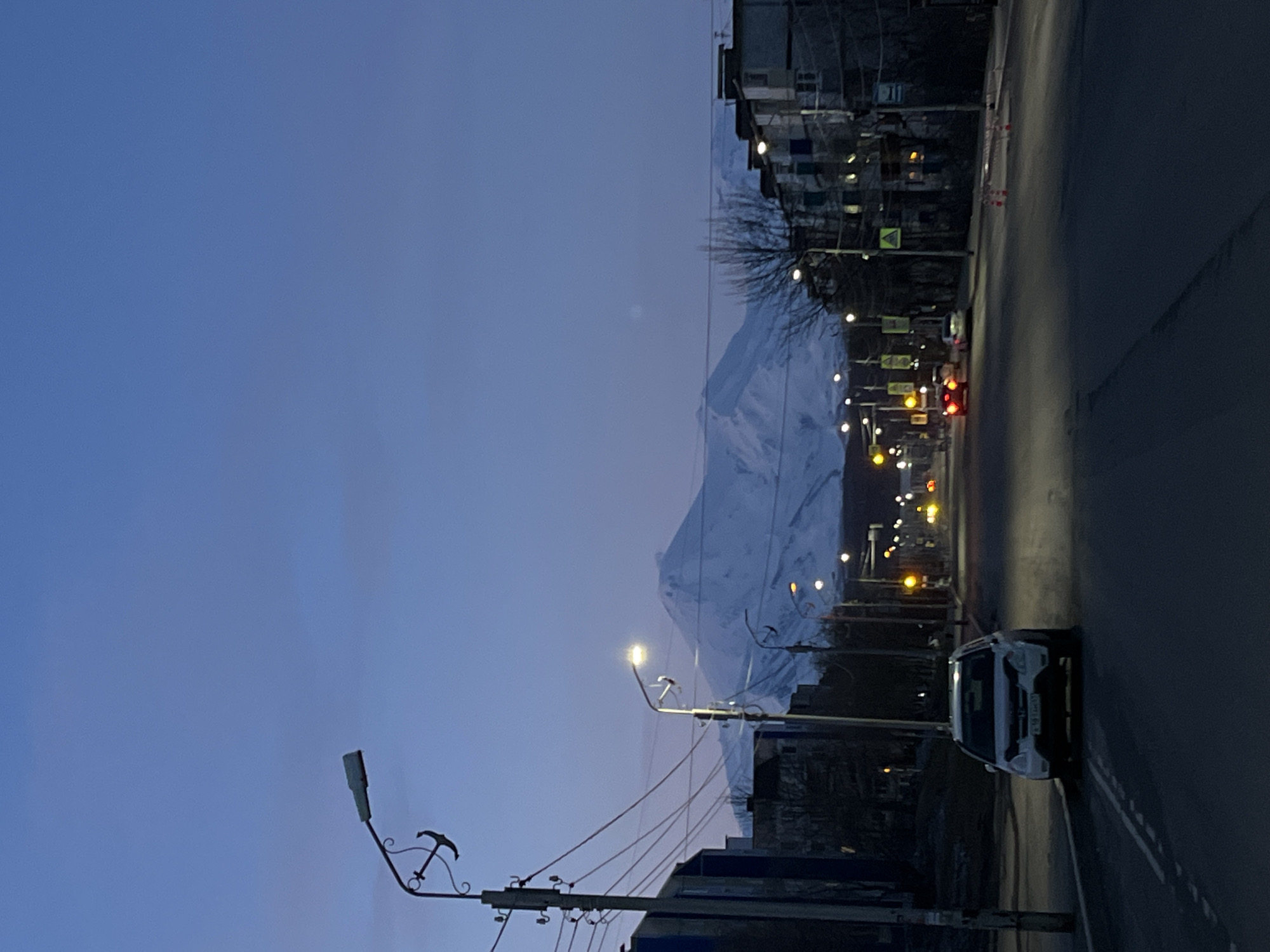 The image shows a street in Petropavlovsk-Kamchatsky, Russia, at dusk or dawn. A snow-covered mountain dominates the background, contrasting with the urban scene. Streetlights illuminate the road and buildings lining the street. A white car is parked on the right side of the road. Power lines are visible, stretching across the foreground. The sky is a gradient of blues and purples.
