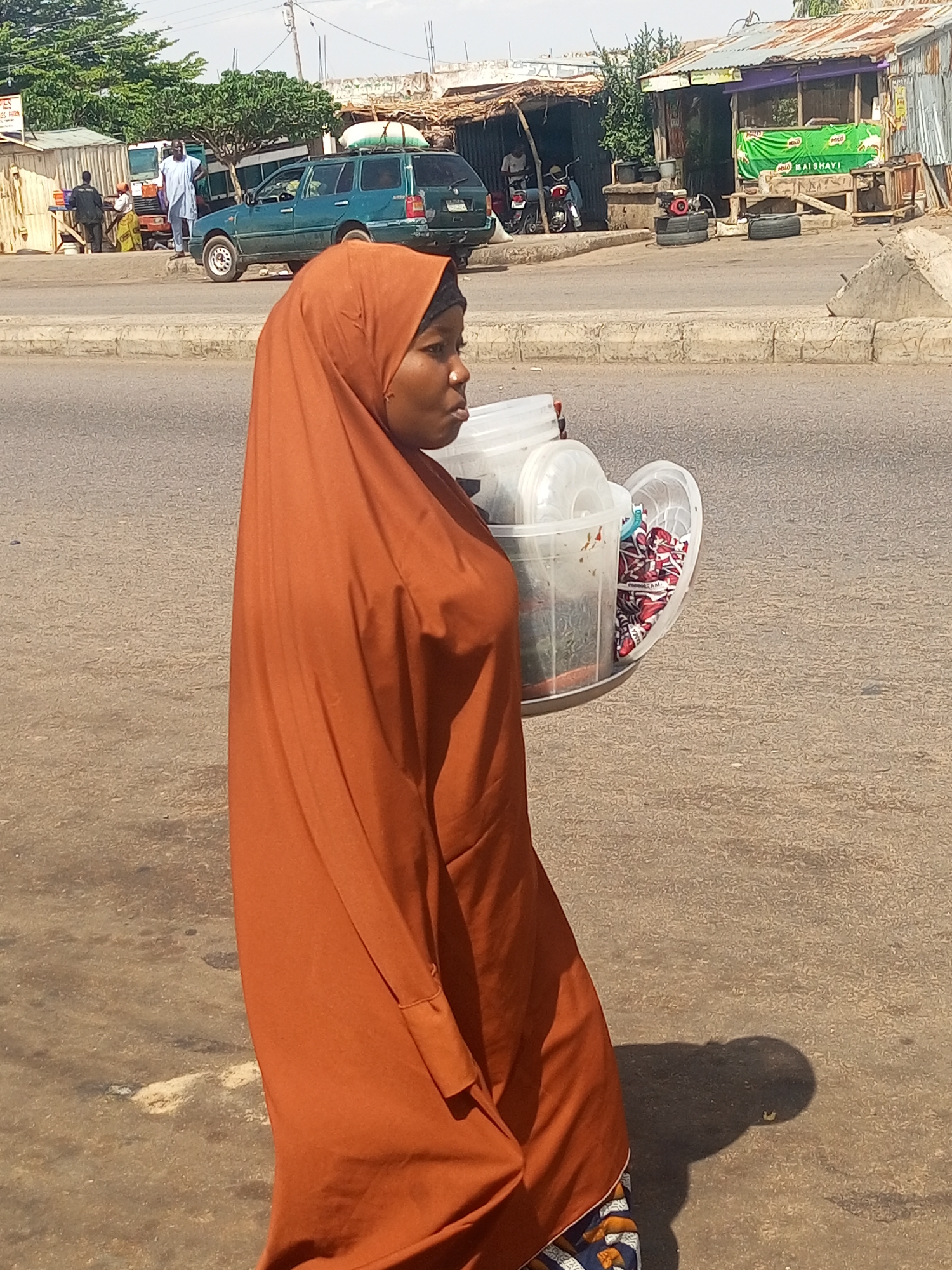 A woman wearing a burnt orange hijab and a patterned long dress walks along a sun-drenched, dusty asphalt road in what appears to be a lively street or market area in Gombe, Nigeria. She is seen in profile, looking slightly to her right, and carries a stack of clear plastic containers, likely buckets or serving dishes, on her right hip. Inside one of the containers, patterned items, possibly fabric or goods for sale, are visible, with text "PRODUCED BY ME" legible on one.
The background reveals a bustling street scene. To the left, a dark green SUV, possibly a Volkswagen model, is parked on the roadside, with items strapped to its roof. Several men are standing nearby, some near a makeshift stall with a metal roof. Further right, more simple structures with corrugated iron roofs line the street, serving as shops or stalls. A motorcycle is parked, and a small red generator rests on two black tires on the ground. A green banner with white text, prominently displaying "MAISHAYI," hangs from one of the stalls.
Strong shadows cast on the road by the woman and other objects indicate bright, overhead sunlight, suggesting it is midday. The overall impression is a snapshot of daily life and commerce in an urban or semi-urban setting.