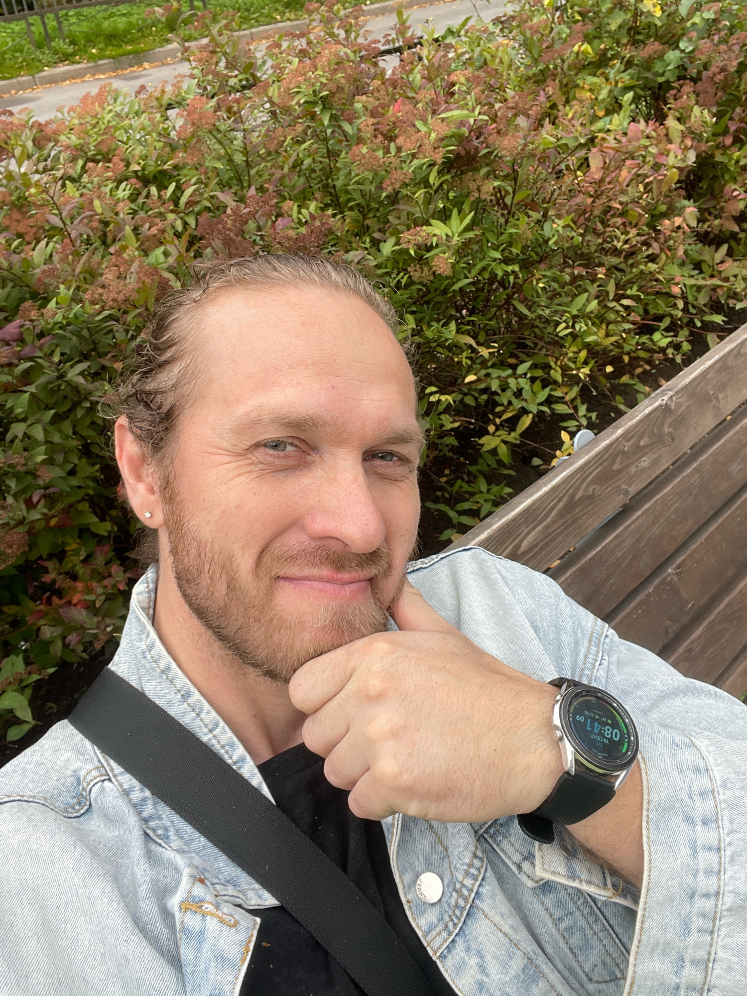 A man with light brown hair pulled back in a bun and a short beard is sitting on a wooden bench in front of a dense bush with green and reddish-brown leaves. He is wearing a light blue denim jacket over a black t-shirt and a black strap is diagonally across his chest. He has a small silver stud earring in his left ear and is wearing a smartwatch on his left wrist. The smartwatch displays "08:41" and other data. He is looking at the camera and touching his chin with his right hand. The background suggests an outdoor urban environment, possibly a park or a street with greenery. The lighting indicates it is daytime.