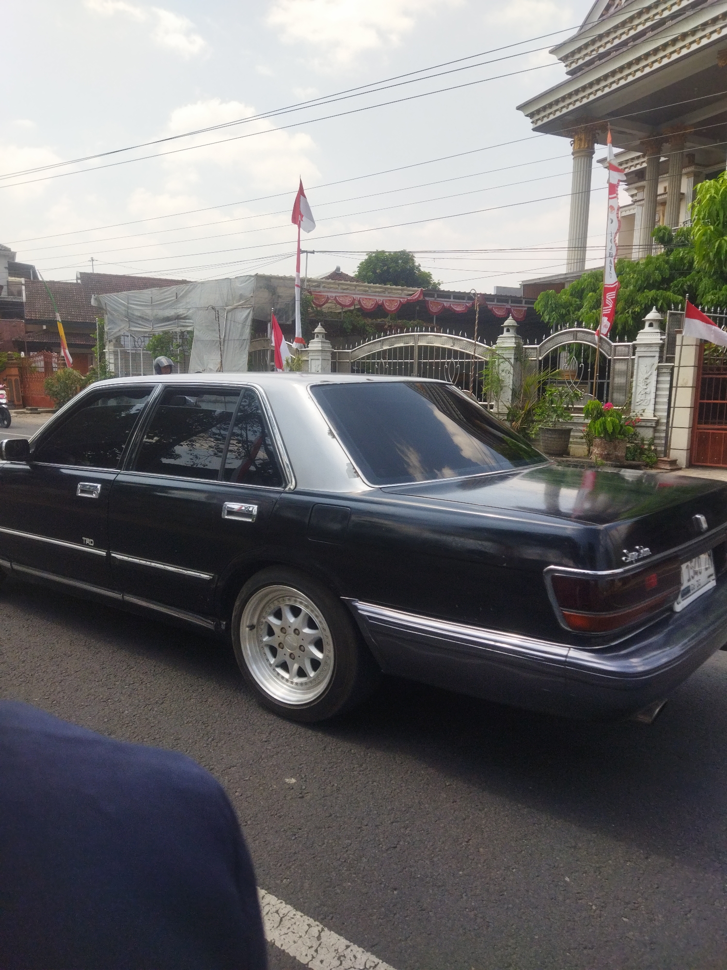 The image shows a black Toyota Crown sedan, possibly from the late 1980s or early 1990s, parked on a street in Nambangan, Indonesia. The car has a silver roof and tinted windows. It is positioned on the right side of the frame, with its rear facing towards the right. The street is asphalt and has a white dashed line on the right side. In the background, there is a large white building with classical columns, a metal fence, and several Indonesian flags displayed. The sky is cloudy. A dark blue object, possibly clothing or a person, is in the foreground, partially obscuring the lower left corner of the image. The overall scene suggests a residential or commercial area decorated for a national celebration.