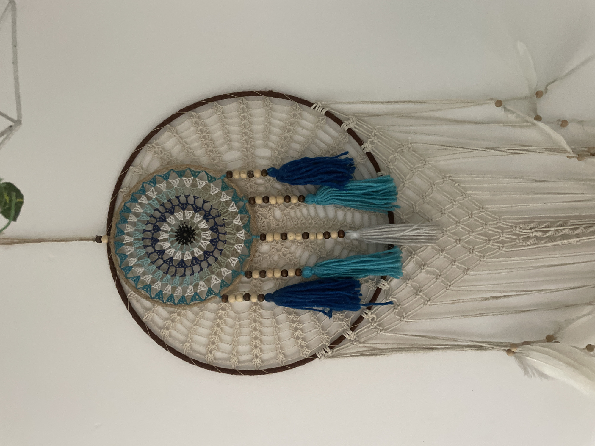 A dreamcatcher is hanging on a plain white wall. The dreamcatcher is circular with a brown outer ring and a woven net inside. In the center of the net is a circular, crocheted design with blue, white, and black threads. Three tassels, two blue and one teal, hang from the bottom of the inner circle, each separated by wooden beads. Below these tassels, a macrame section hangs down, from which several long, off-white strings descend. White feathers are attached to some of these strings near the bottom. The image does not provide any information about the location within Mumbai, India.