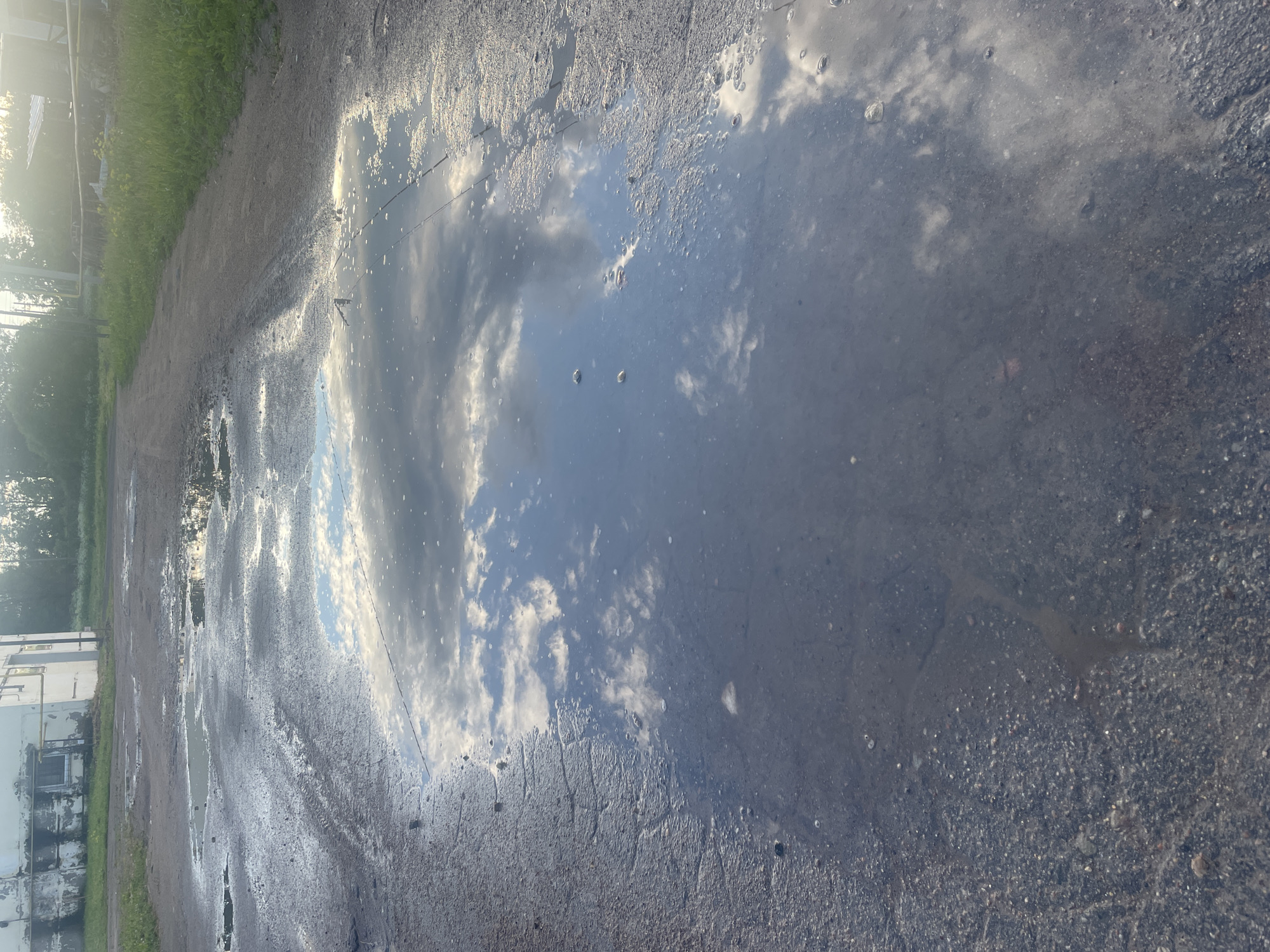 The image shows a cracked asphalt surface with multiple puddles of water. The largest puddle in the foreground reflects a blue sky with white, puffy clouds and sunlight. To the left, in the background, is a weathered white building with some visible piping. The environment appears to be an outdoor area, possibly a yard or industrial space, with vegetation and a dirt path or road visible further in the background. The overall impression is one of a somewhat neglected urban or industrial landscape after rain.