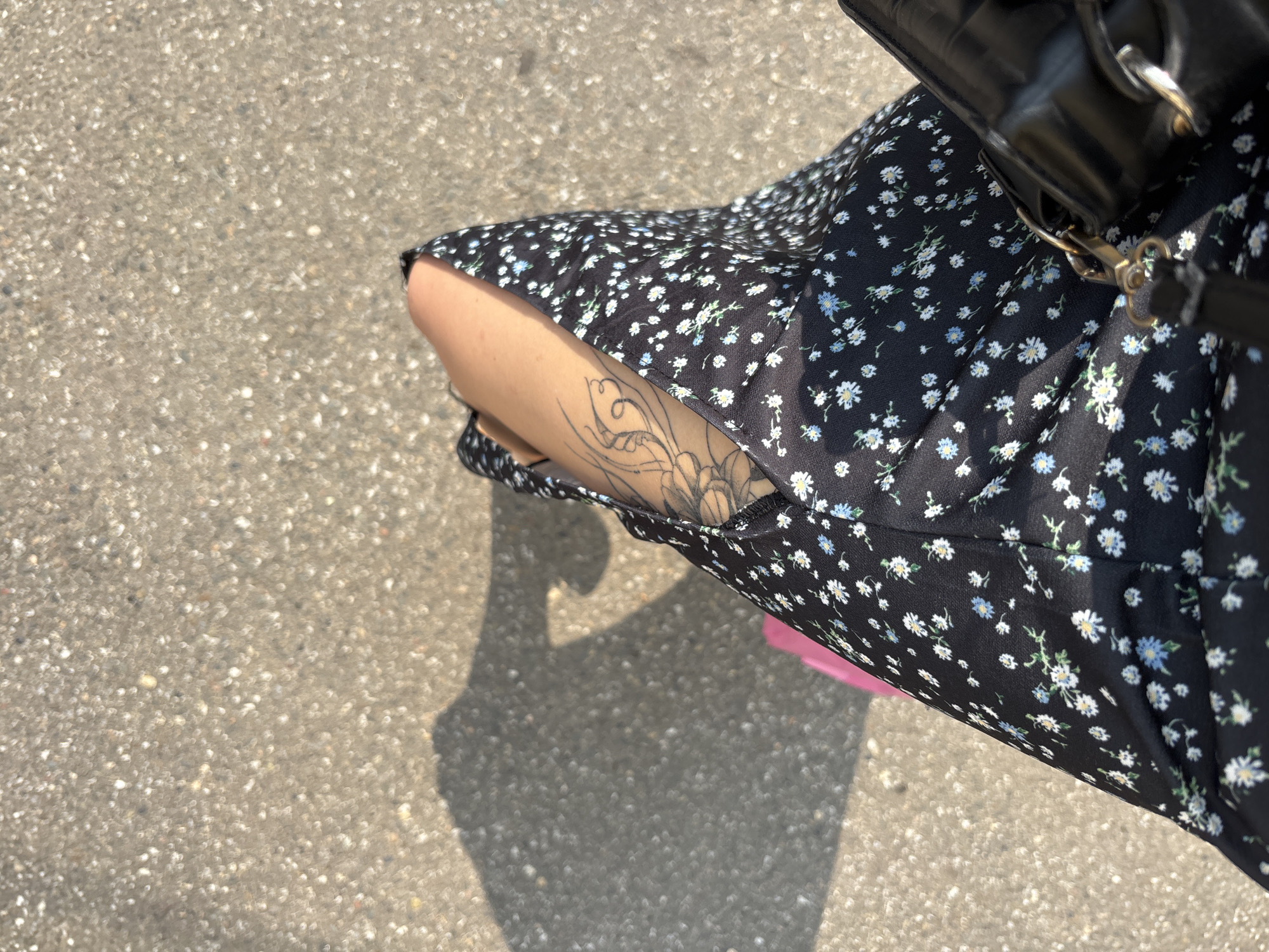 The image shows a close-up view of a person's legs and arm, resting on a paved surface, likely asphalt. The person is wearing a black dress with a small floral print of white and blue flowers. A tattoo is visible on the person's arm, featuring intricate lines and shading. A black handbag with a quilted texture and gold hardware rests beside the person. A shadow is cast on the ground, suggesting it is a sunny day. Part of a pink object, possibly a skateboard or accessory, is visible near the person's legs. The environment appears to be an outdoor urban setting in Novokuznetsk, Russia, given the paved ground and the context provided.