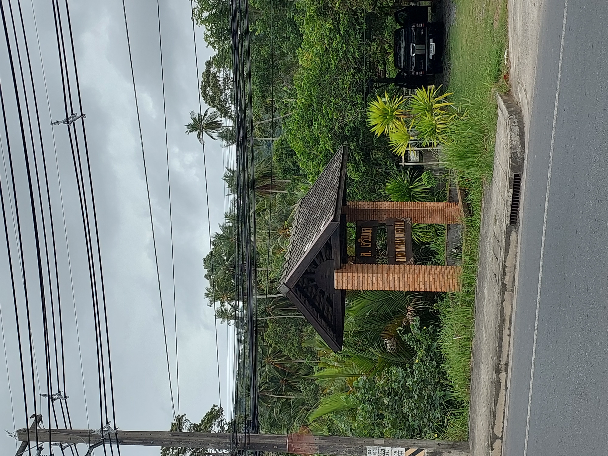 A black pickup truck is parked in an open area on the right side of the image, with its doors open. In front of the truck, there is lush green vegetation and tall grass. To the left of the truck, a sign is mounted on brick pillars and topped with a wooden roof. The sign has text in Thai and English that reads "BANG MAKHAM ROENTHAI." A sidewalk runs along the bottom of the image, separating the road from the vegetated area. The road is paved and has a white line marking the lane. A utility pole with multiple black wires strung across it stands on the left side of the image. The background is filled with dense green trees, including palm trees, and a cloudy sky. The scene appears to be in a tropical or subtropical environment.