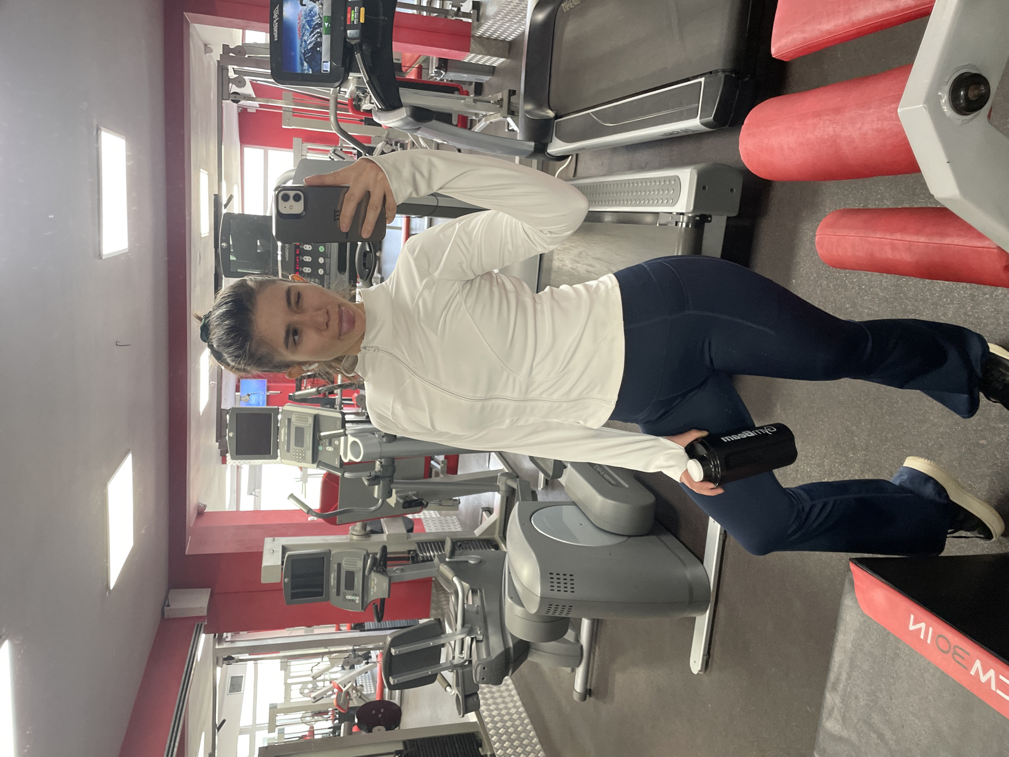 A young woman is taking a selfie in a gym. She is wearing a white long-sleeved top and dark blue leggings and is holding a black water bottle. The gym has various exercise machines, including treadmills and elliptical trainers. The gym's walls are painted red and white. Based on the presence of weight machines bearing the "NEWB01N" logo, the gym is likely located in Sofia, Bulgaria, as this logo is associated with a fitness center in that city.