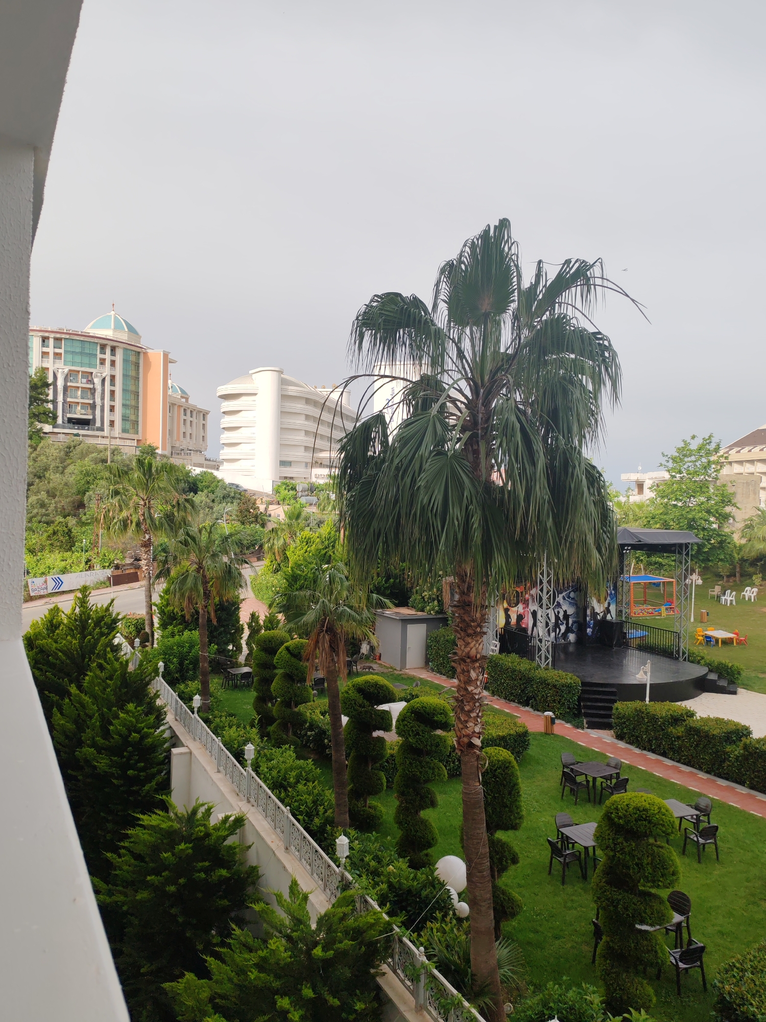 The image shows an outdoor stage in a landscaped area, likely a hotel garden, in Alanya, Turkey. The stage is set against a backdrop of stylized graffiti figures and has black stairs leading up to it. A colorful play area is situated next to the stage. There are scattered plastic chairs and tables on the grass, arranged for guests to sit. The landscape includes well-maintained shrubs and several palm trees. In the background, there are modern buildings, one with a distinctive turquoise dome. The scene is viewed from an elevated position, possibly from a balcony, as part of a white railing is visible on the left side of the image. The sky is overcast.
