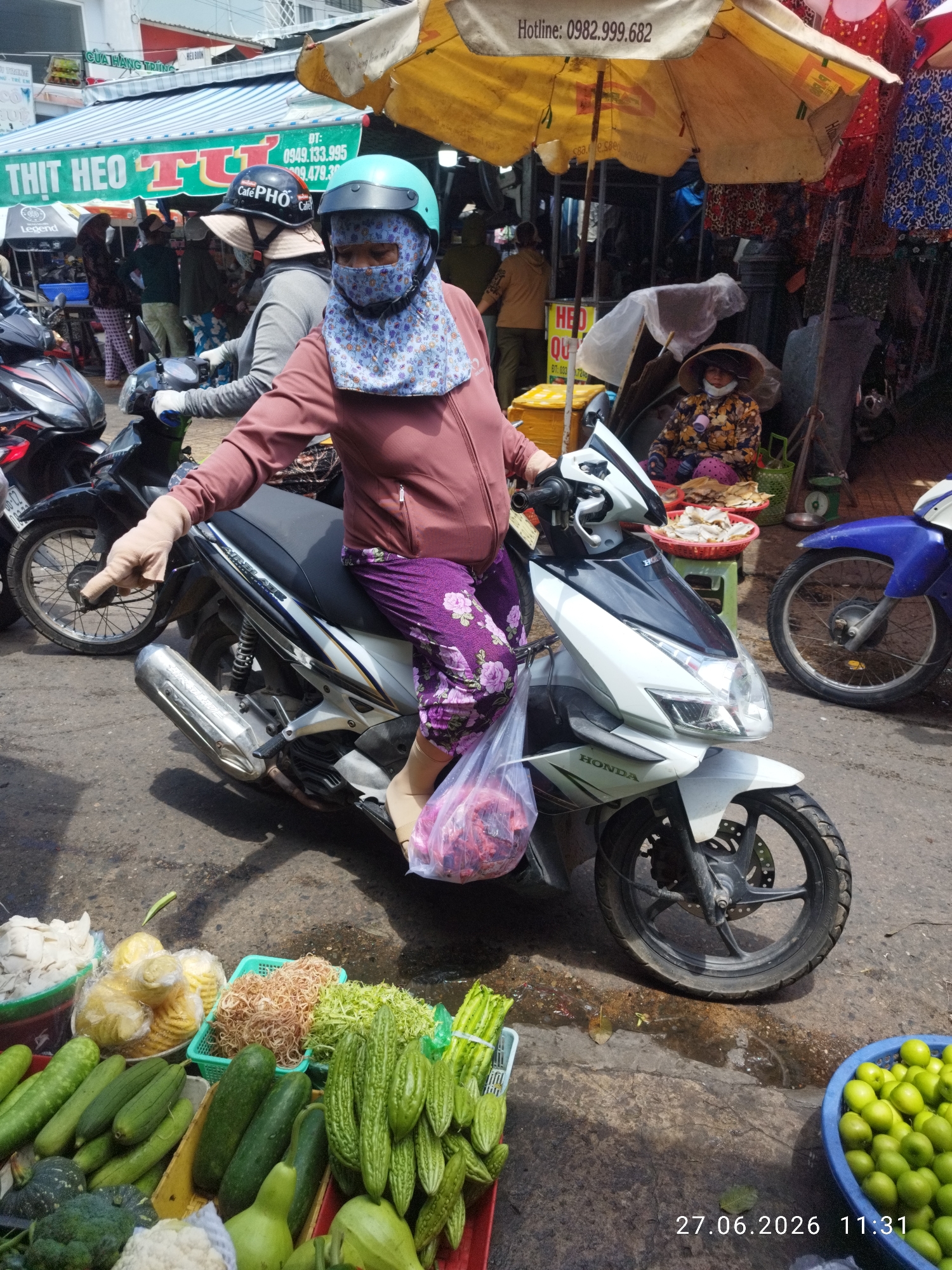 A woman wearing a pink jacket, purple floral pants, and a light blue patterned headscarf and mask rides a white Honda motorcycle in a market. She wears a light blue helmet and light beige gloves. A clear plastic bag filled with red items hangs from the handlebars. The motorcycle's front wheel is turned to the left, and its side stand is down. In the background, several other motorcycles are parked, and market stalls display various goods, including vegetables, fish, and clothing. One stall has a yellow umbrella. The ground is paved with asphalt and appears wet in some areas. The scene is set outdoors in a busy market environment, likely in Phan Thiết, Vietnam, as indicated by the presence of fresh produce and local vendors.