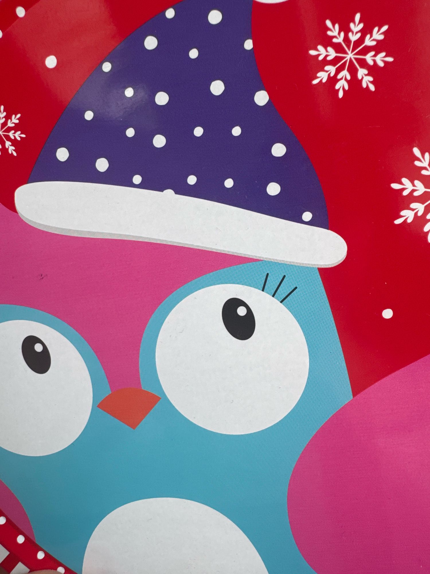 The image displays a close-up of a brightly colored, cartoonish illustration of an owl wearing a Santa hat, set against a red background with white snowflakes. The owl is predominantly blue with large white eyes, a pink chest, and a small orange beak. It is wearing a purple Santa hat adorned with white polka dots and a white brim. The background features white snowflake designs and white polka dots scattered across the red surface. The scene is graphic and appears to be part of a festive decoration or packaging. There are no elements in the image that indicate a specific location in the city of Katowice, Poland.