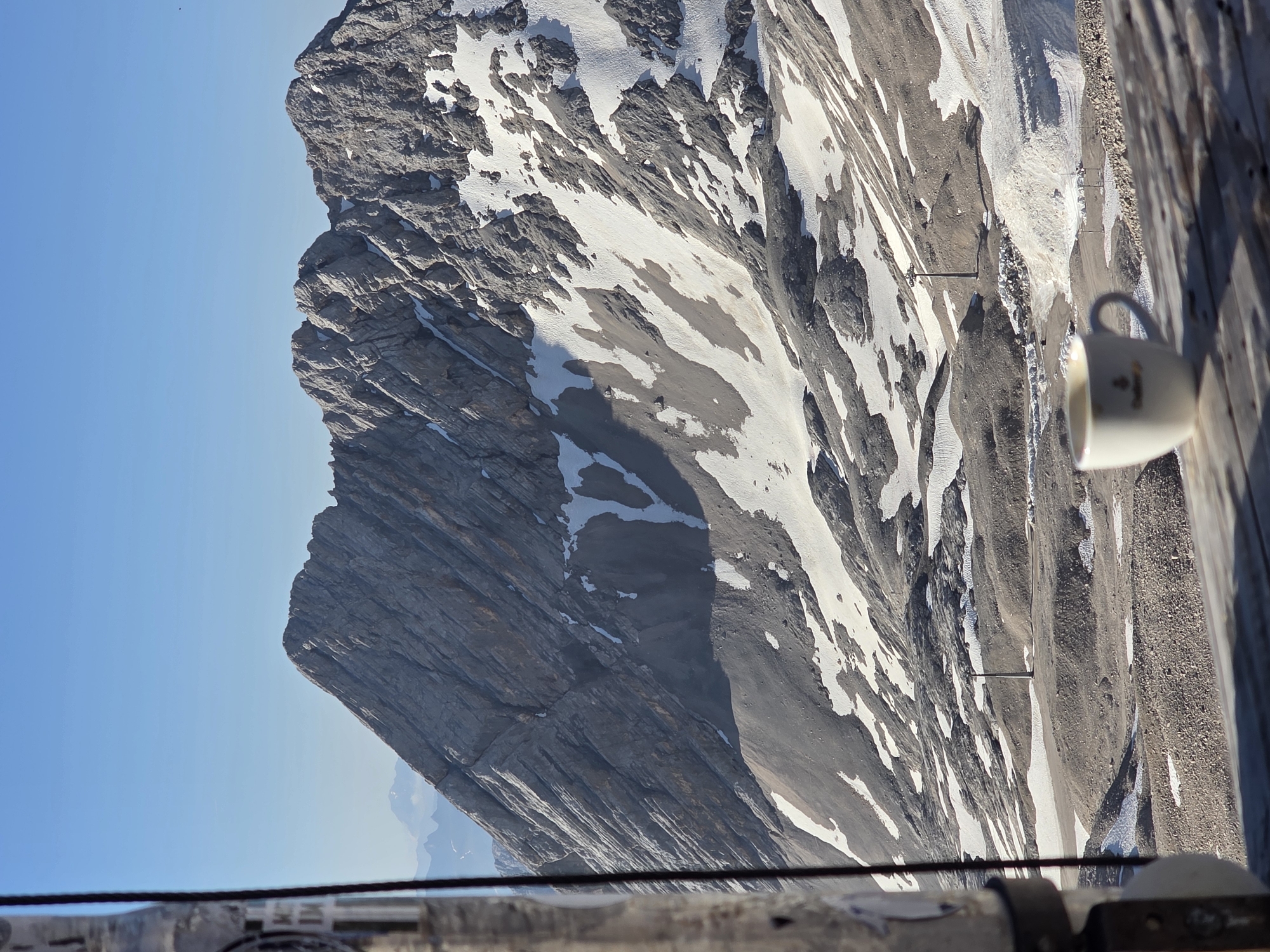 The image depicts a stunning mountain landscape with a clear blue sky. The mountain is characterized by steep, rocky cliffs with patches of snow and scree. The texture of the rock layers is prominent, showcasing the geological formations. In the foreground, a wooden surface with planks suggests a balcony or observation deck. A white mug with a subtle logo is placed on this surface, adding a touch of human presence to the rugged natural scene. The perspective is from a high vantage point, looking down and across the vast mountain range. The lighting suggests it is daytime, likely with bright sunshine given the shadows and clear sky. The scene evokes a sense of awe and tranquility, possibly a moment of rest and appreciation of the mountain scenery. The location is identified as Garmisch-Partenkirchen, Germany, a region known for its alpine beauty. There is a faint logo on the mug, but it is not clearly legible.