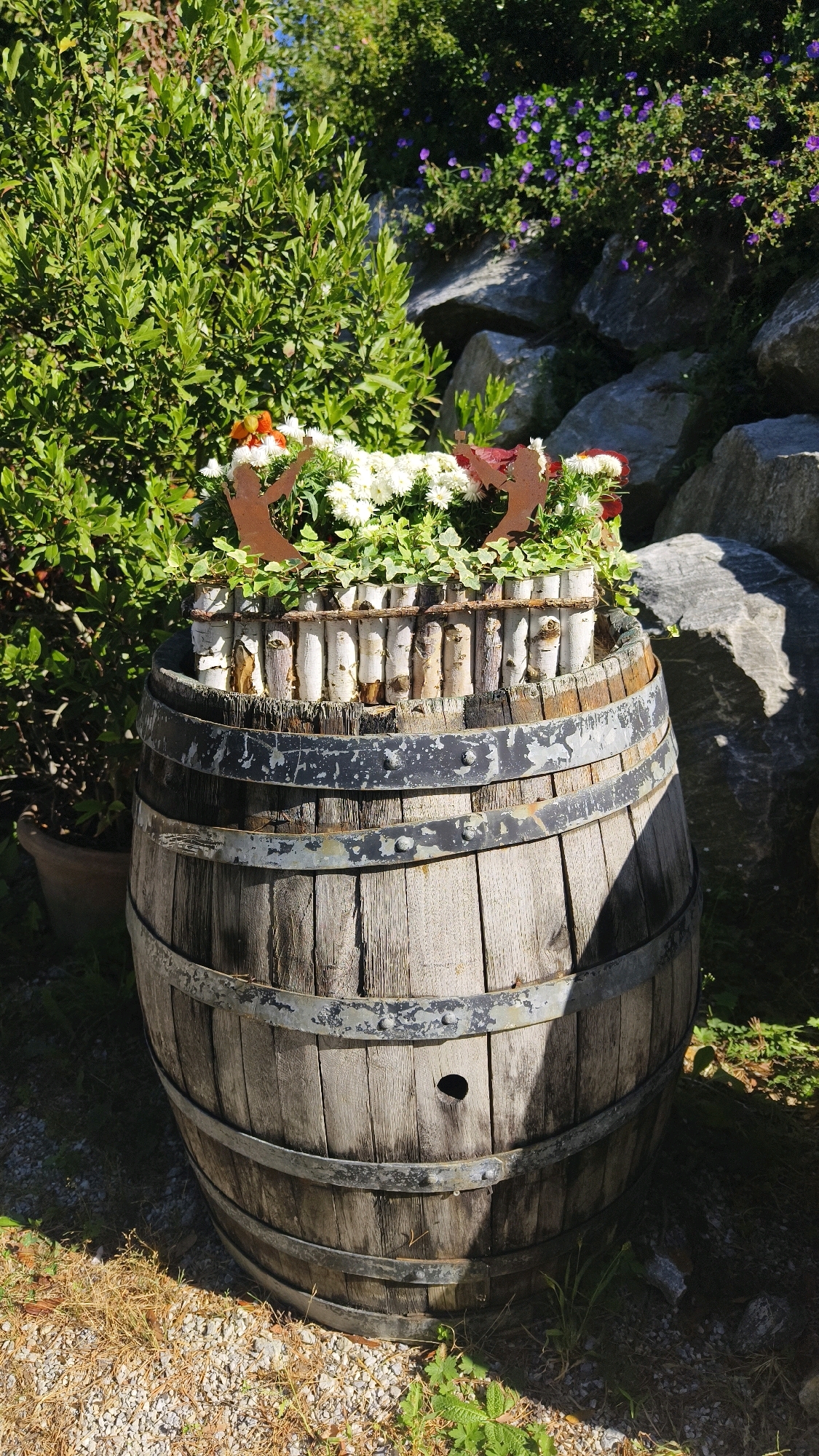 The image depicts a decorative arrangement featuring a rustic wooden barrel as a planter, situated outdoors in what appears to be a garden or natural setting. The barrel is filled with white and orange flowers, interspersed with green foliage and some decorative wooden elements around the rim. Two silhouette figures, appearing to be made of metal, are visible amongst the flowers. Behind the barrel, lush green bushes and large rocks are prominent, with smaller purple flowers visible in the background. The ground is covered with gravel and dry grass. The overall scene suggests an outdoor garden decoration. While the image is identified as being in Langenlois, Austria, there are no specific landmarks or features that pinpoint an exact city location within Langenlois.