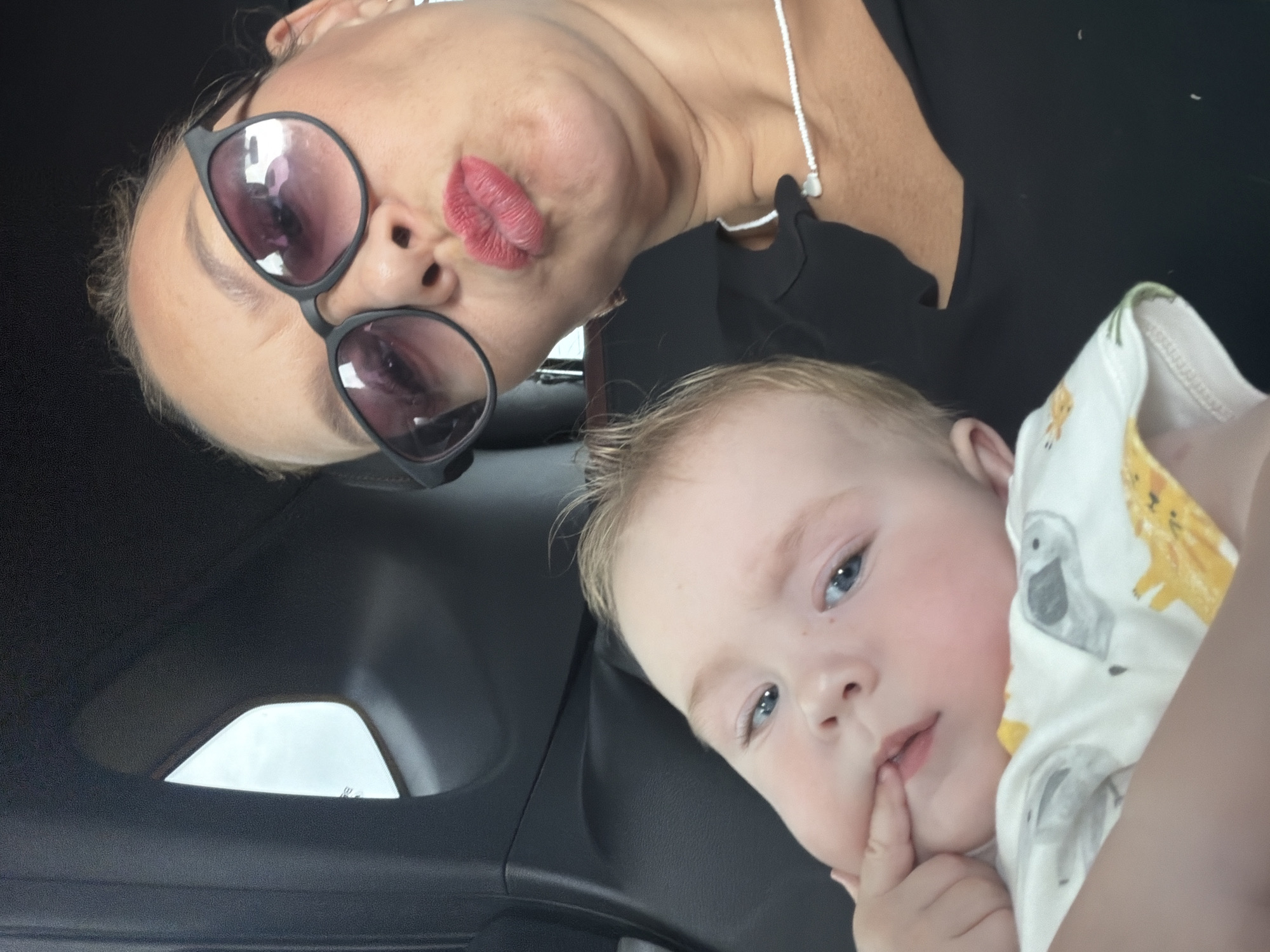 In an indoor setting, possibly a car, a woman and a baby are captured in a selfie. The woman, wearing sunglasses and lipstick, puckers her lips for the camera. Her hair is pulled back, and she has on a black top with a delicate beaded necklace. The baby, with blue eyes and fair hair, is held in the woman's arms, with one finger resting on its lips. The baby is dressed in a white onesie with a colorful animal print. The background shows the interior of a vehicle, with a window providing a glimpse of the sky. The overall ambiance suggests a casual, personal moment. The location is Povarenki, Russia.