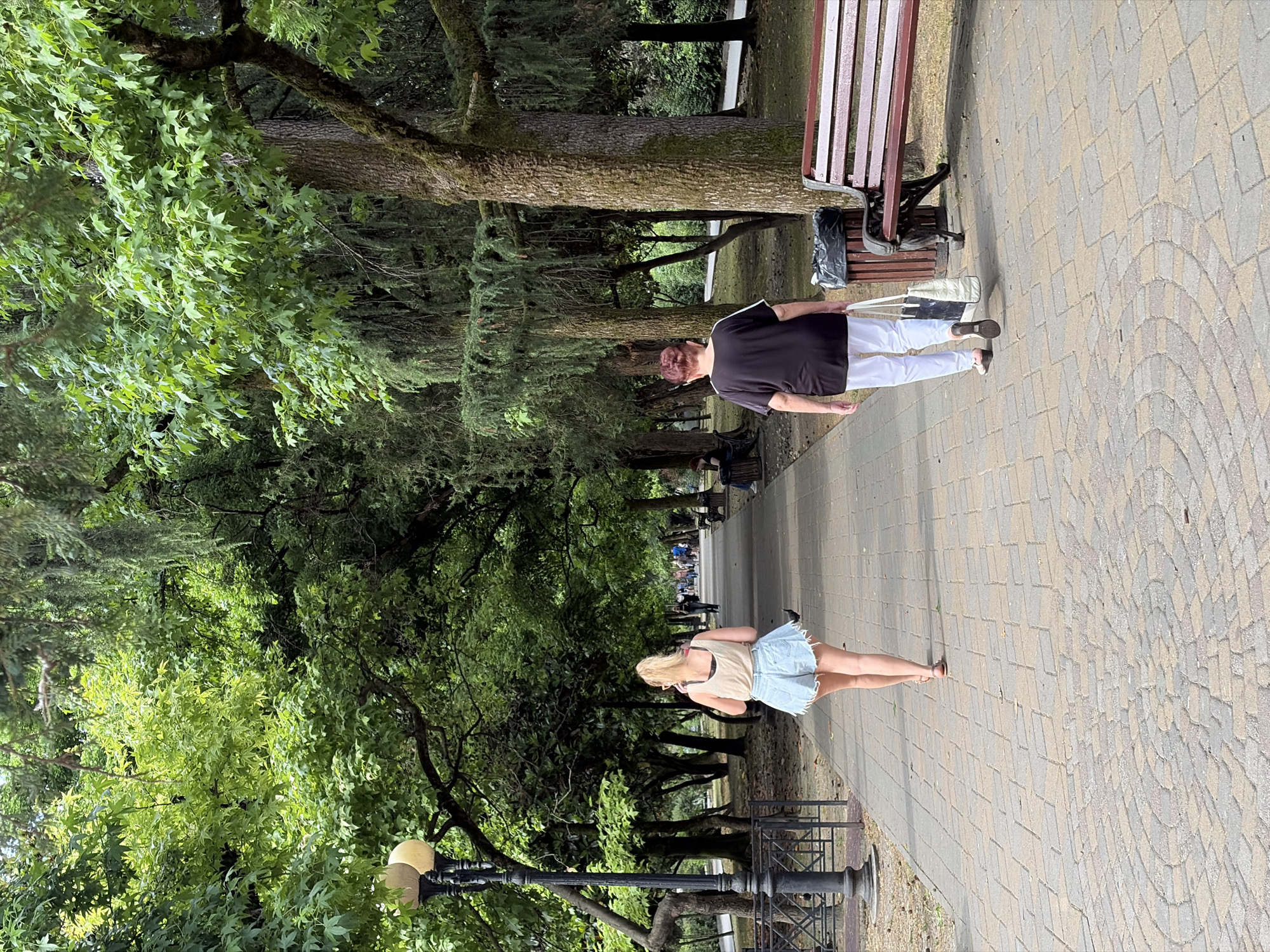A scene in a park in Sochi, Russia, shows two women walking away from the camera on a paved walkway. The woman in the foreground on the left has blonde hair and is wearing a light-colored tank top and denim shorts. The woman on the right has red hair and is wearing a black t-shirt with white stripes and white pants, carrying a tote bag. The walkway is lined with trees and benches. A lamppost is visible on the left side of the frame. In the background, other people are walking and sitting on benches. The environment is green and lush with mature trees.
