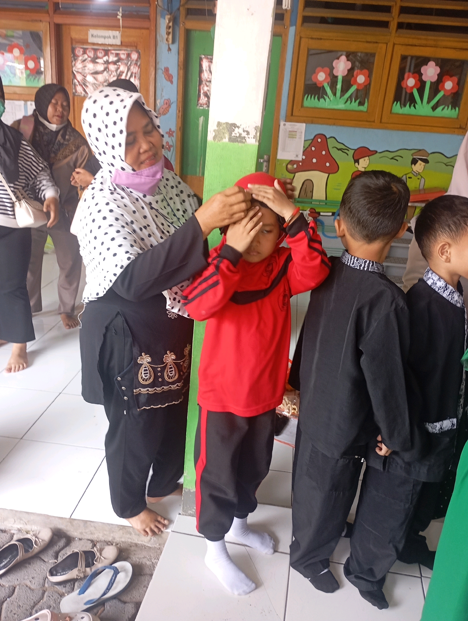 In Babelan, Indonesia, a woman wearing a black and white polka dot hijab and a purple face mask adjusts the red headscarf of a young child. The child is wearing a red long-sleeved shirt with black stripes and black pants. Behind them, two boys are dressed in black uniforms. The environment appears to be a classroom or a communal space, with colorful drawings of flowers and a mushroom house on a mural covering the wall. A wooden door with a sign that reads "Kelompok B1" (Group B1) is visible in the background. Shoes are neatly placed on the floor near the entrance.