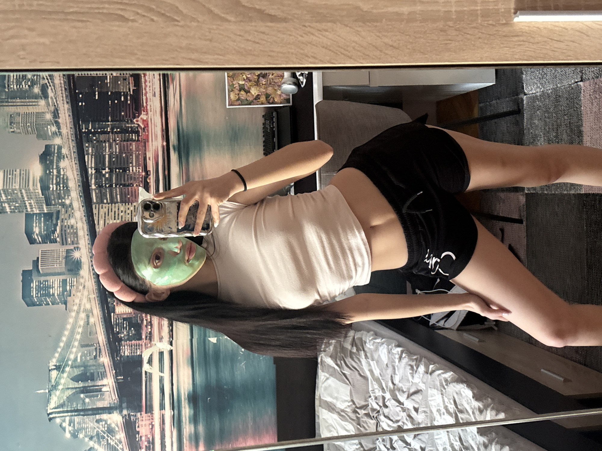 A young woman with long black hair and a pink headband is posing for a selfie in front of a mirror. She has a green facial mask on. She is wearing a white t-shirt and black shorts. The background shows a cityscape, possibly a poster or reflection in the mirror, reminiscent of a cityscape with bridges. A bed is visible below, with crumpled white sheets. The reflection also shows a chair and a carpeted floor. There is no indication of Sofia, Bulgaria in the image.