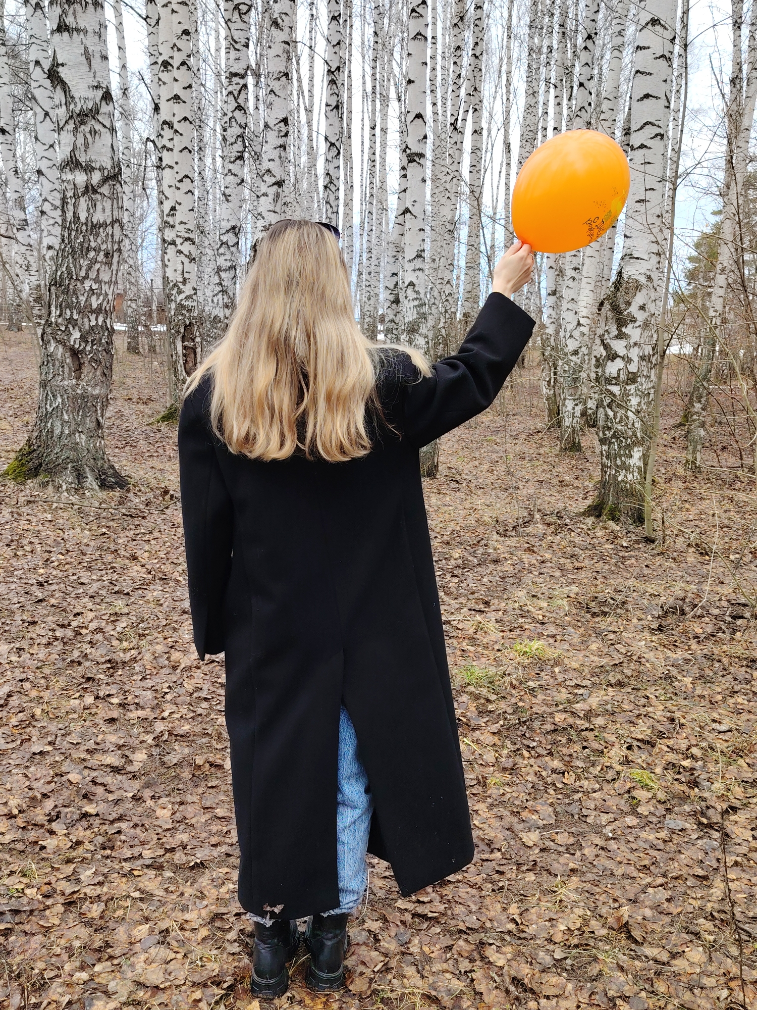 The image shows a woman standing in a birch forest. The ground is covered with fallen leaves. The woman has long blonde hair and is wearing a black coat, blue jeans, and black boots. She is holding an orange balloon in her right hand. The location is in the city of Chaikovsky, Russia.