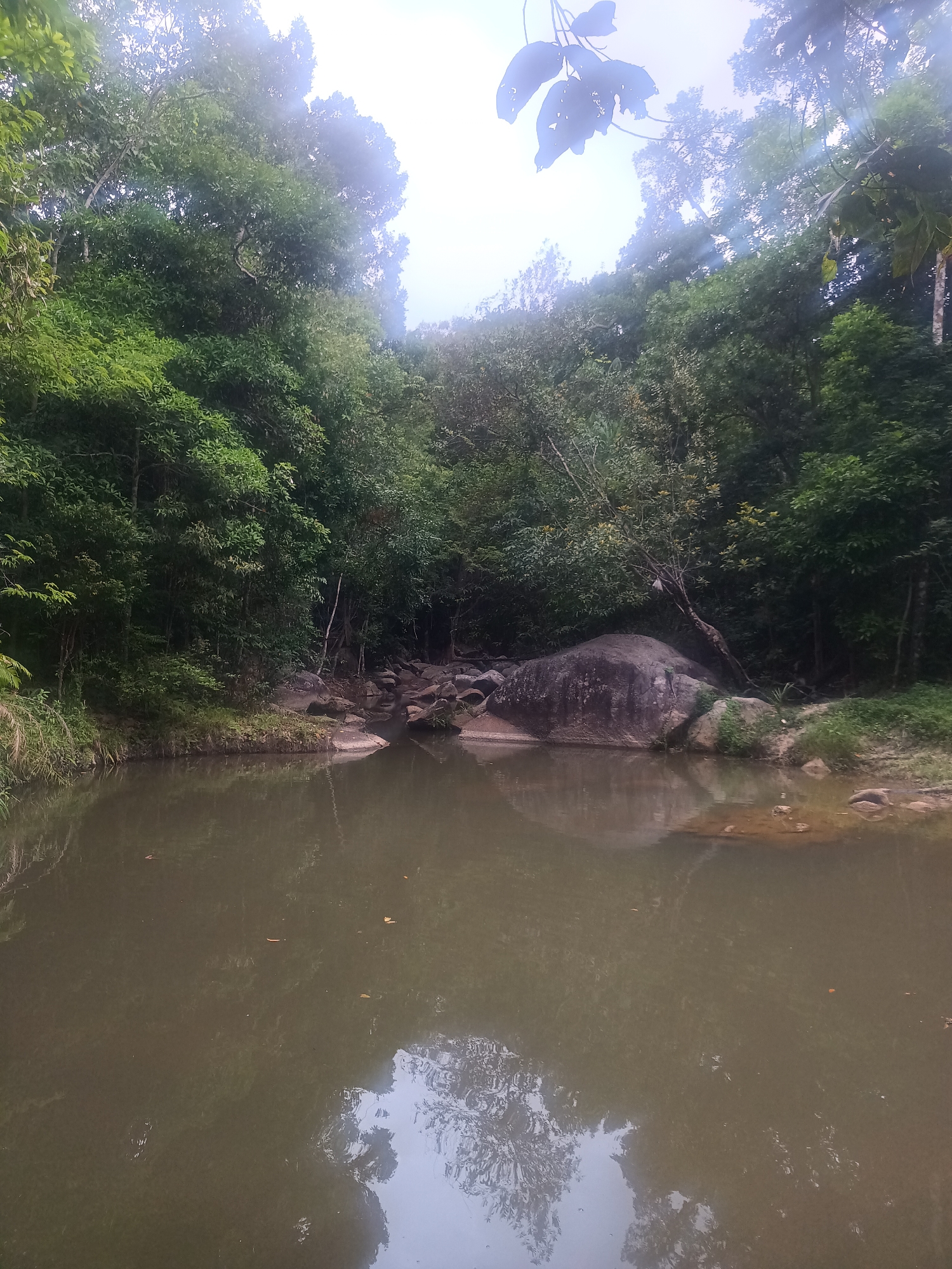 The image depicts a small, calm body of water bordered by lush green vegetation. The location is Baan Thong Nang, Thailand. The water is murky, reflecting the sky and trees above. The surrounding area consists of trees and rocks, suggesting a natural, possibly forested environment. No people are visible in the frame.